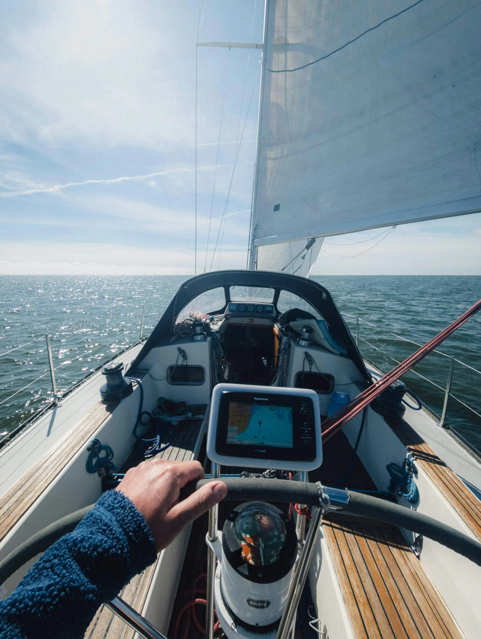 A person steering a sailboat on open water with a view of the boat's dashboard and navigation equipment, sails, and a calm ocean horizon.
