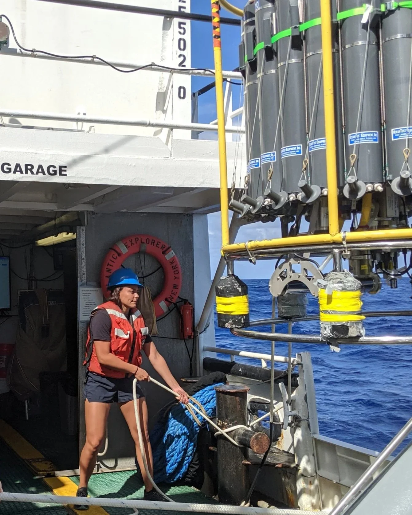 A woman wearing a blue helmet and a life vest on a boat, handling a coiled blue rope, with a background of the ocean and equipment on the boat.