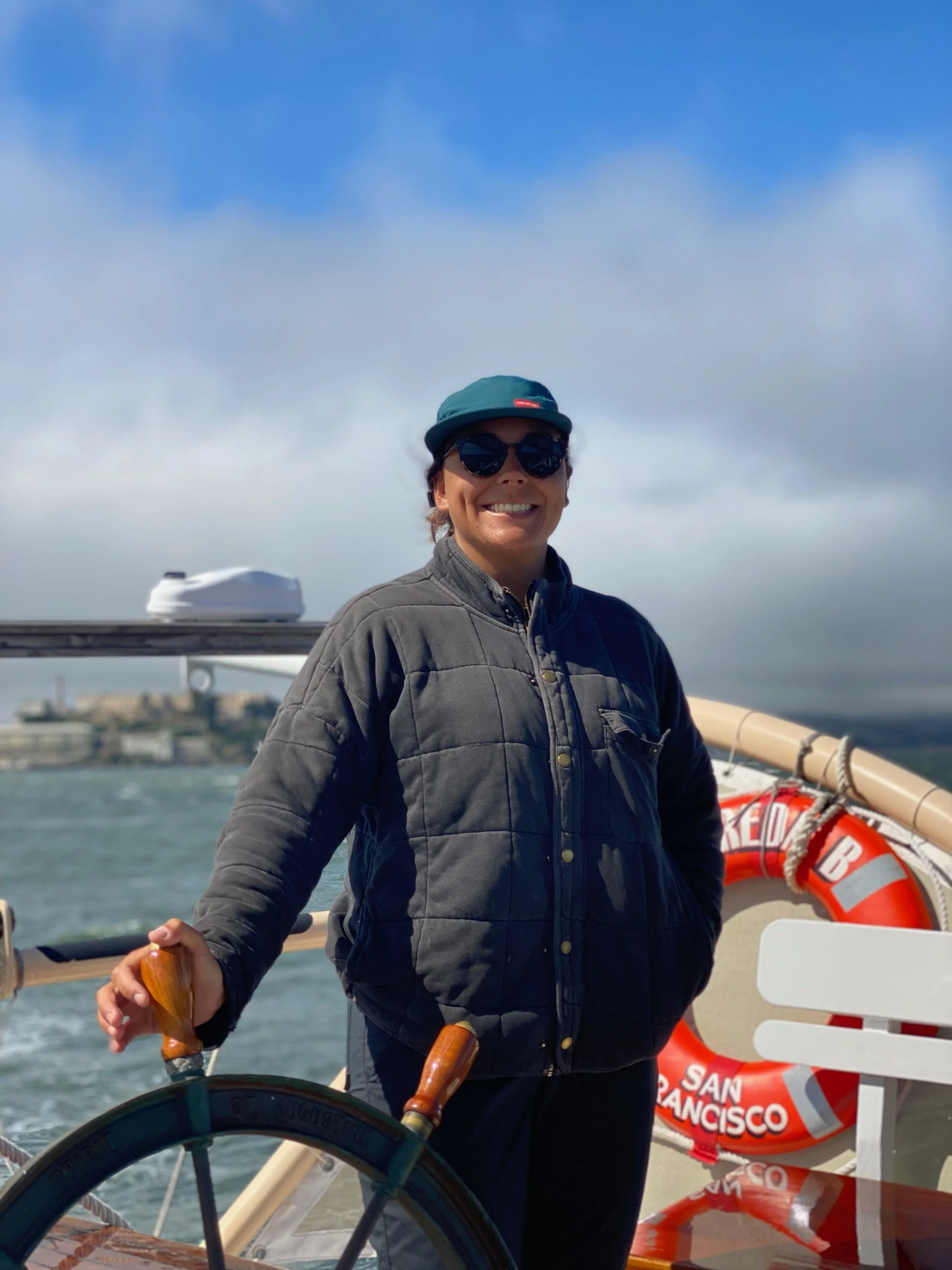 A smiling woman wearing sunglasses, a green cap, a gray jacket, and holding a wooden steering wheel on a boat, with a life ring reading 'San Francisco' and water in the background.