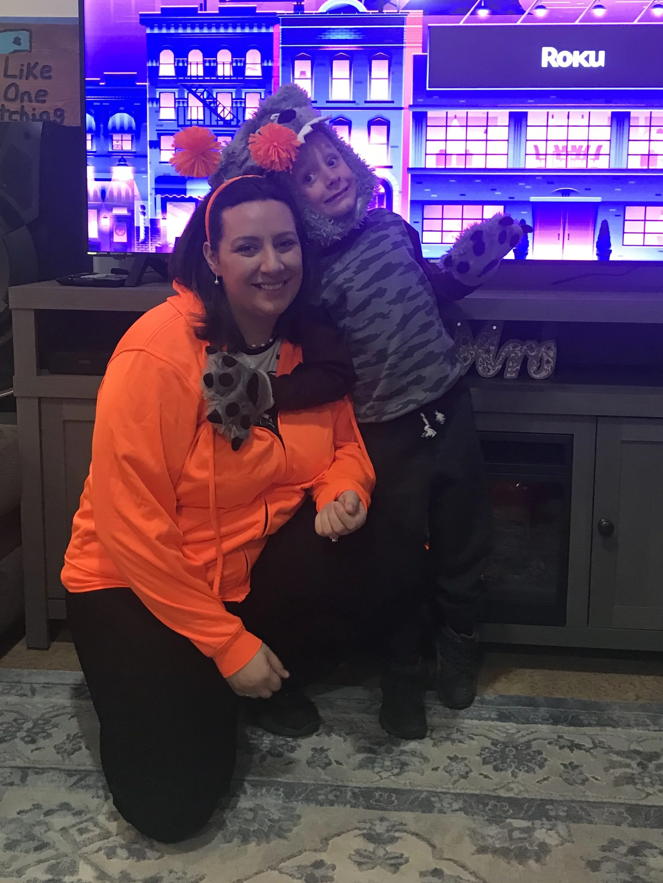A woman dressed in an orange jacket and black pants kneels beside a child wearing a wolf costume with a furry hood and paw gloves. They are indoors in front of a TV screen that displays the Roku logo, with a brightly lit animated cityscape backdrop. There is a patterned rug on the floor and a small table with electronic equipment behind them.