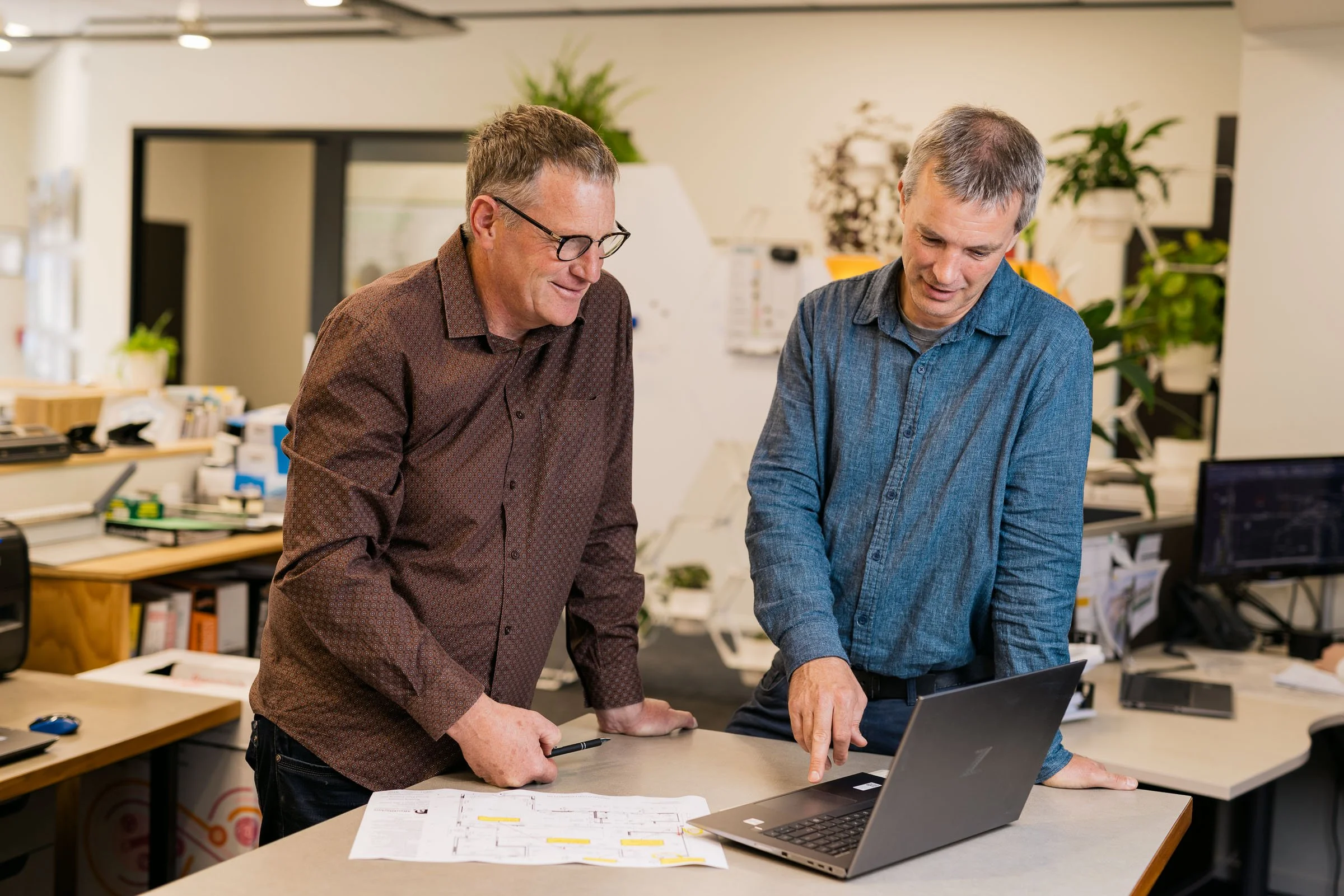 Two men are standing at a table in an office, looking at a laptop and discussing documents. One man is wearing glasses and a patterned shirt, and the other is wearing a blue button-up shirt. There are office supplies and plants in the background.