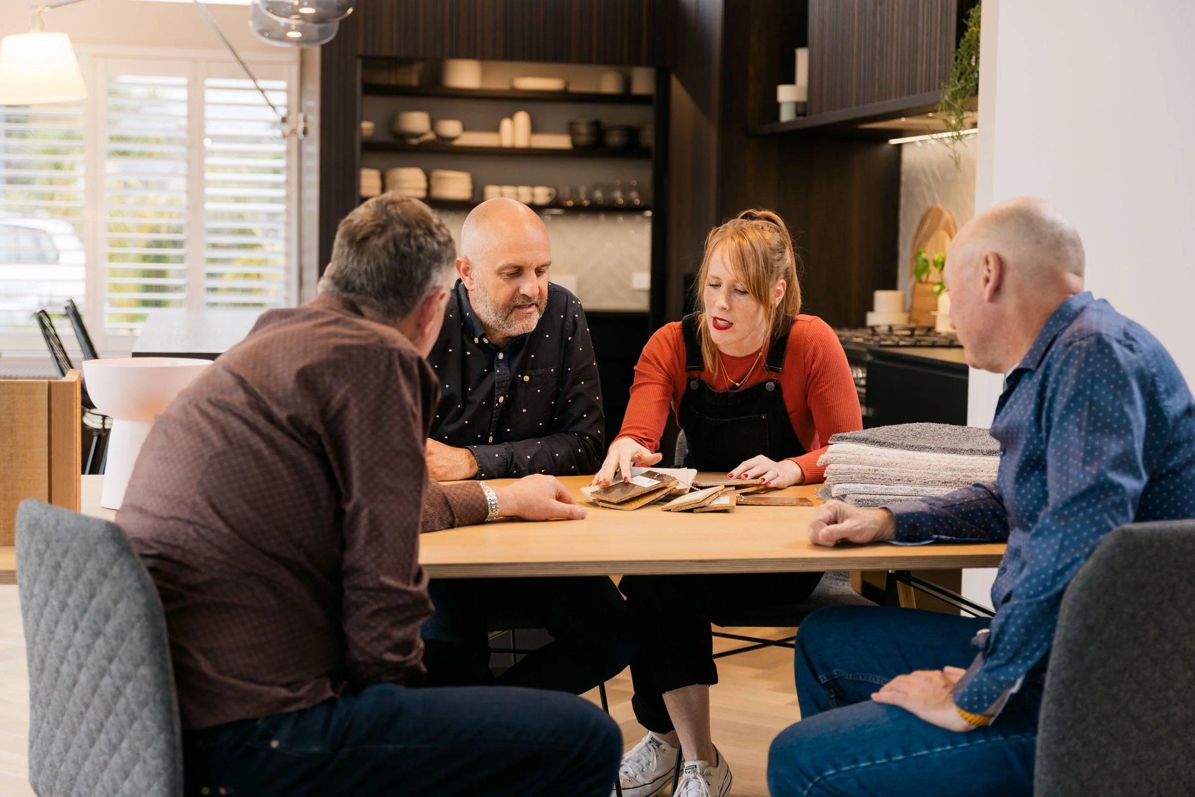 Four people sitting around a wooden table discussing fabric samples in a kitchen-like space.