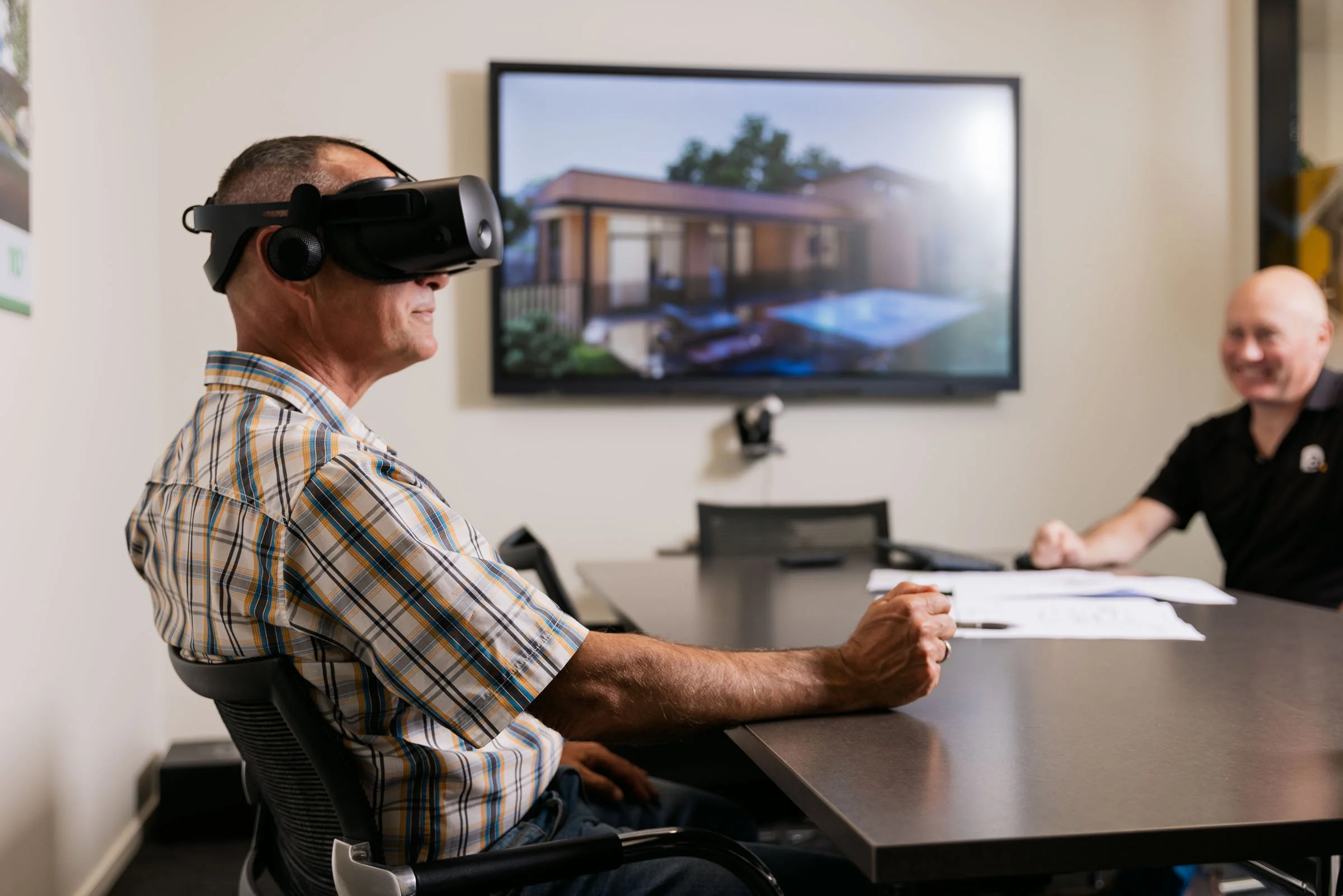 A man wearing a virtual reality headset sitting at a table during a meeting, with a woman smiling in the background in a conference room.