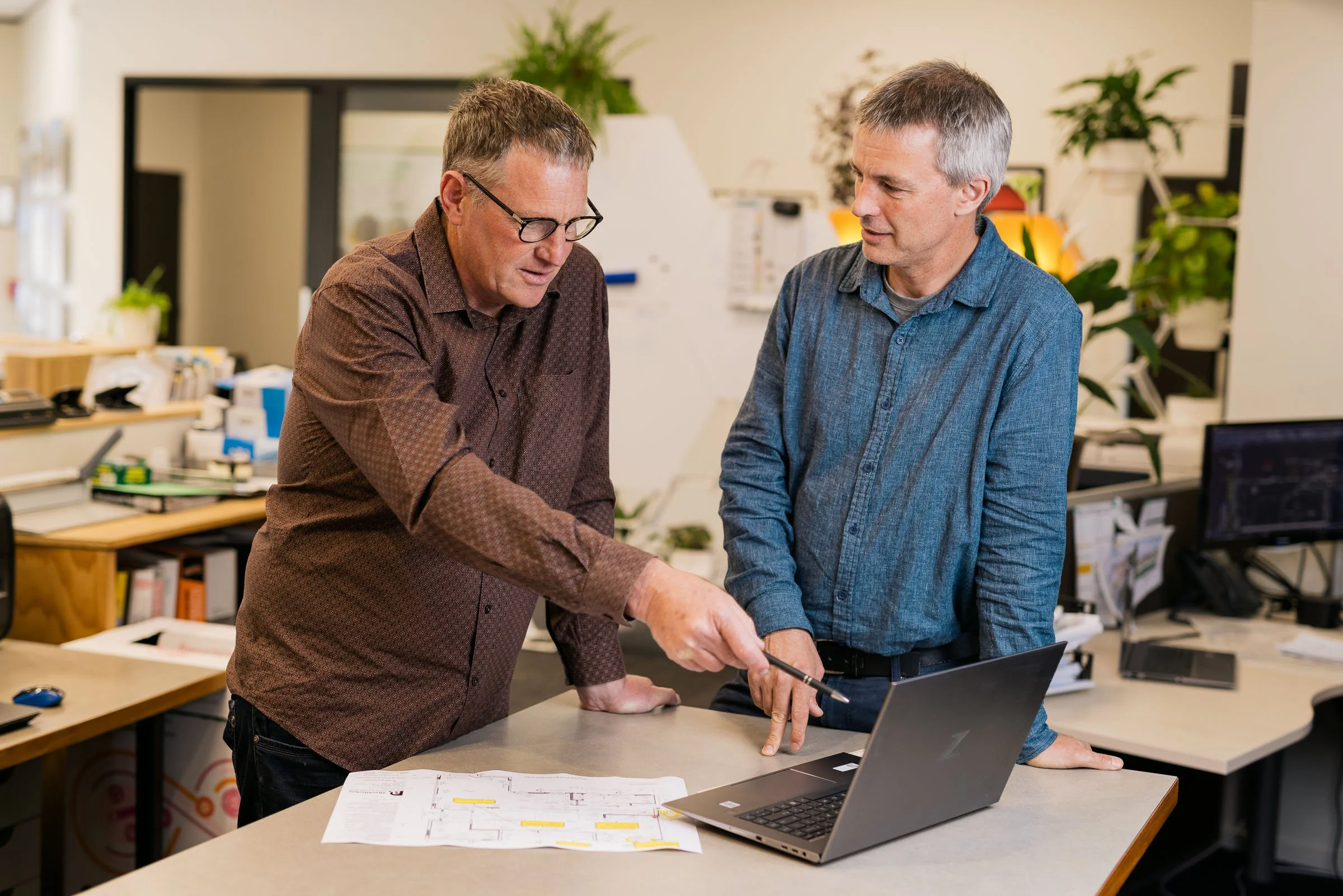 Two men in a discussion over a laptop and papers in an office setting, with shelves and plants in the background.