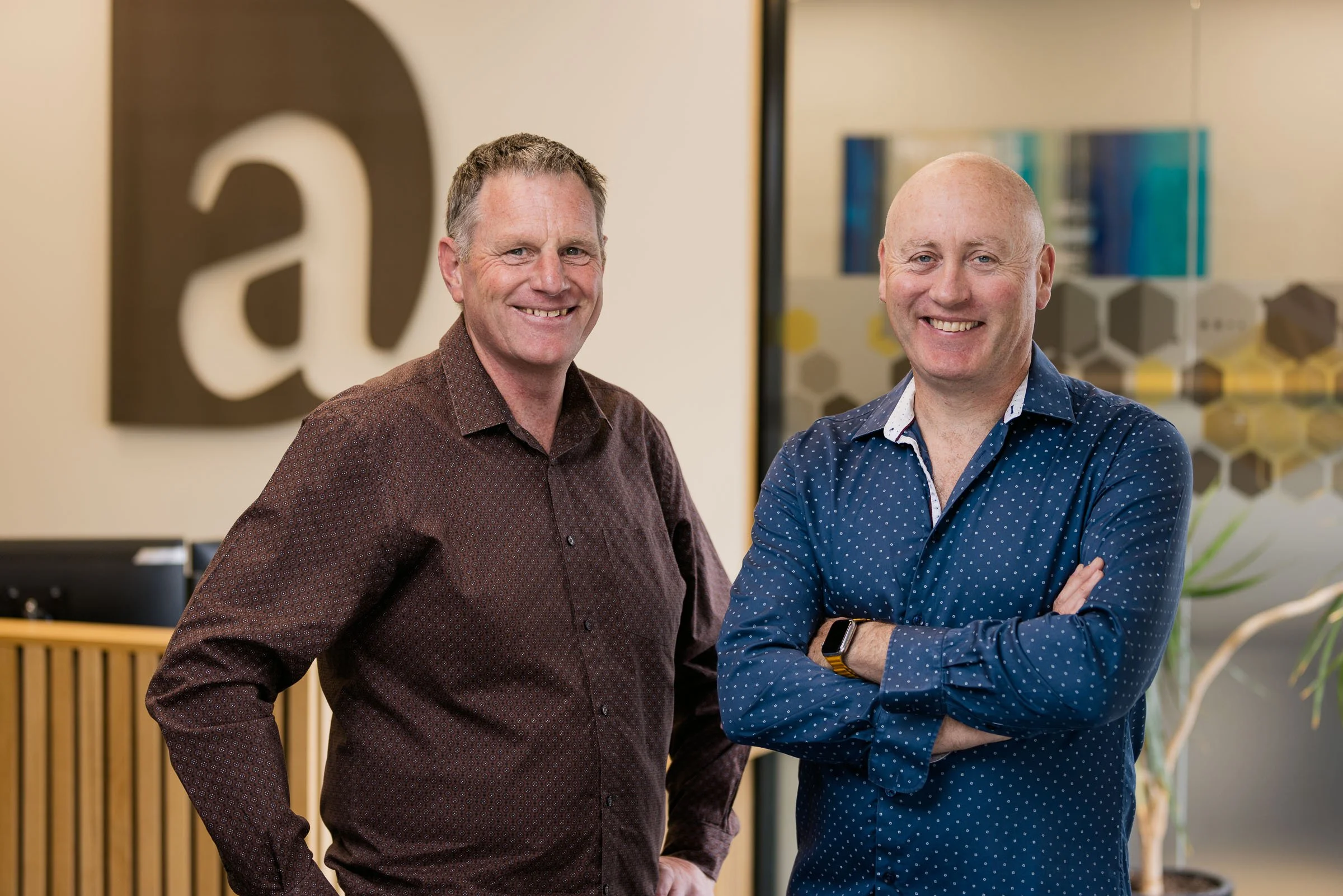 Two smiling men standing indoors, one with hands in pockets and the other with arms crossed. Background includes office decor and wall art.
