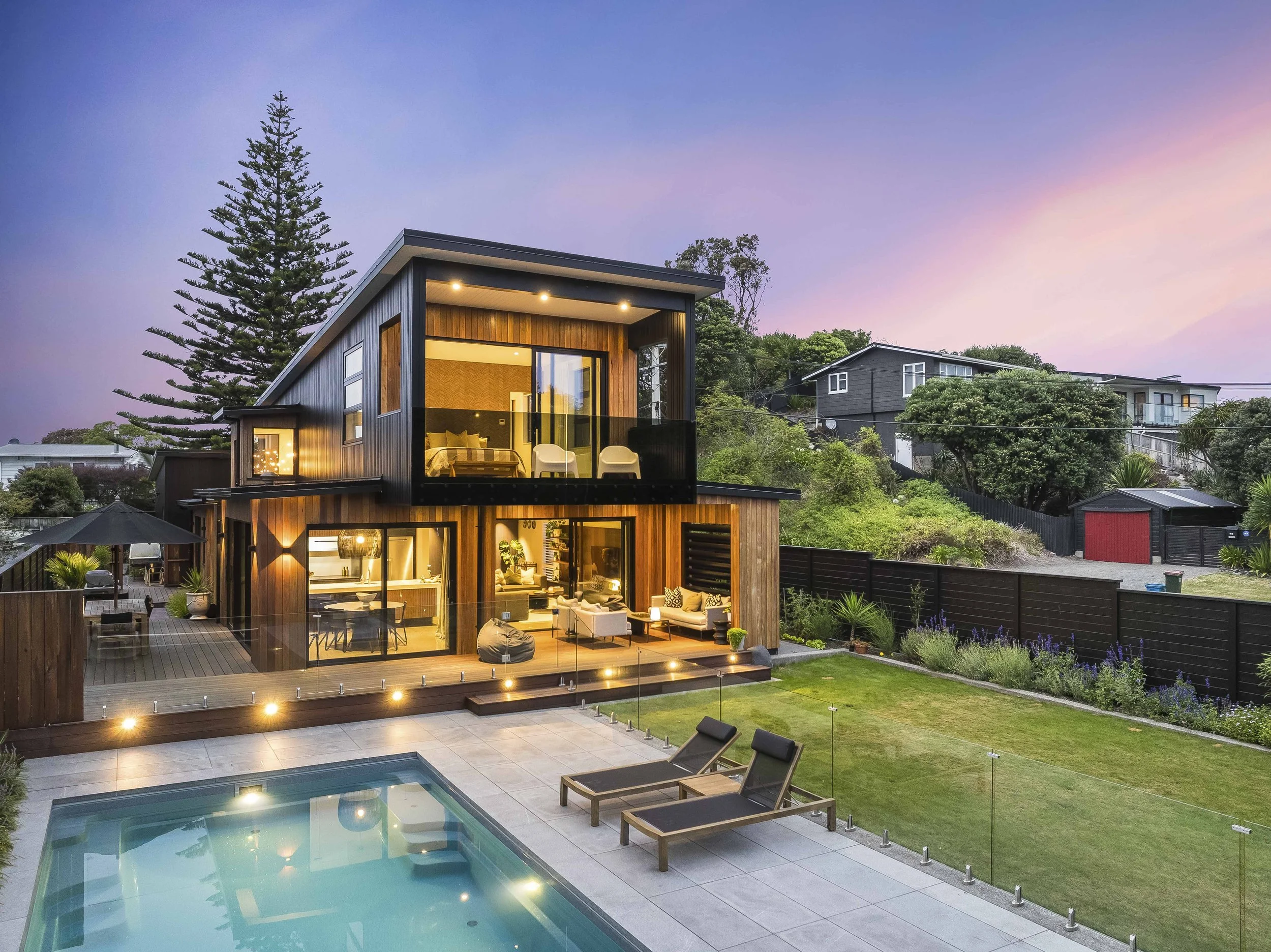 Modern two-story house with wooden exterior and large glass sliding doors, illuminated at dusk. A backyard with a swimming pool, two lounge chairs, and a grassy area with plants and shrubs. Surrounding trees and neighboring houses are visible in the background.
