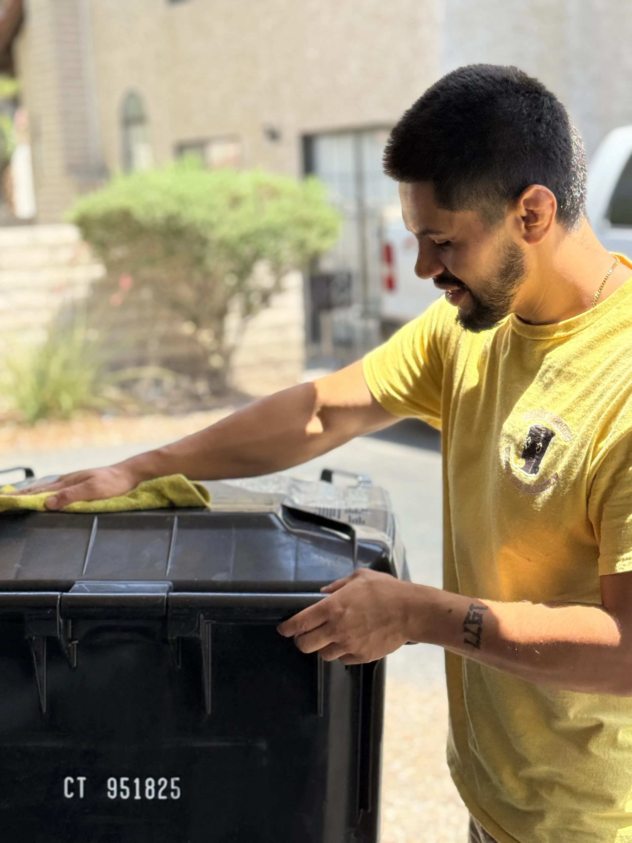 A young man with short black hair, a beard, and tattoos on his arms is cleaning a black grill with a yellow cloth outdoors. He is wearing a yellow T-shirt and appears to be focused on his task.