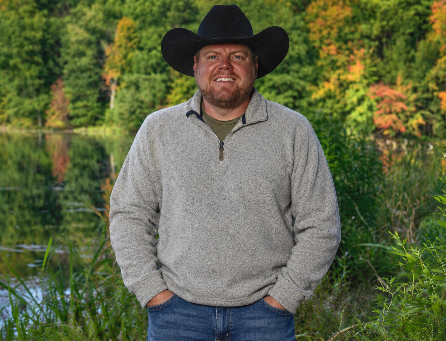 A man wearing a black cowboy hat and a light gray quarter zip sweater poses outdoors near a body of water, with lush green trees in the background.