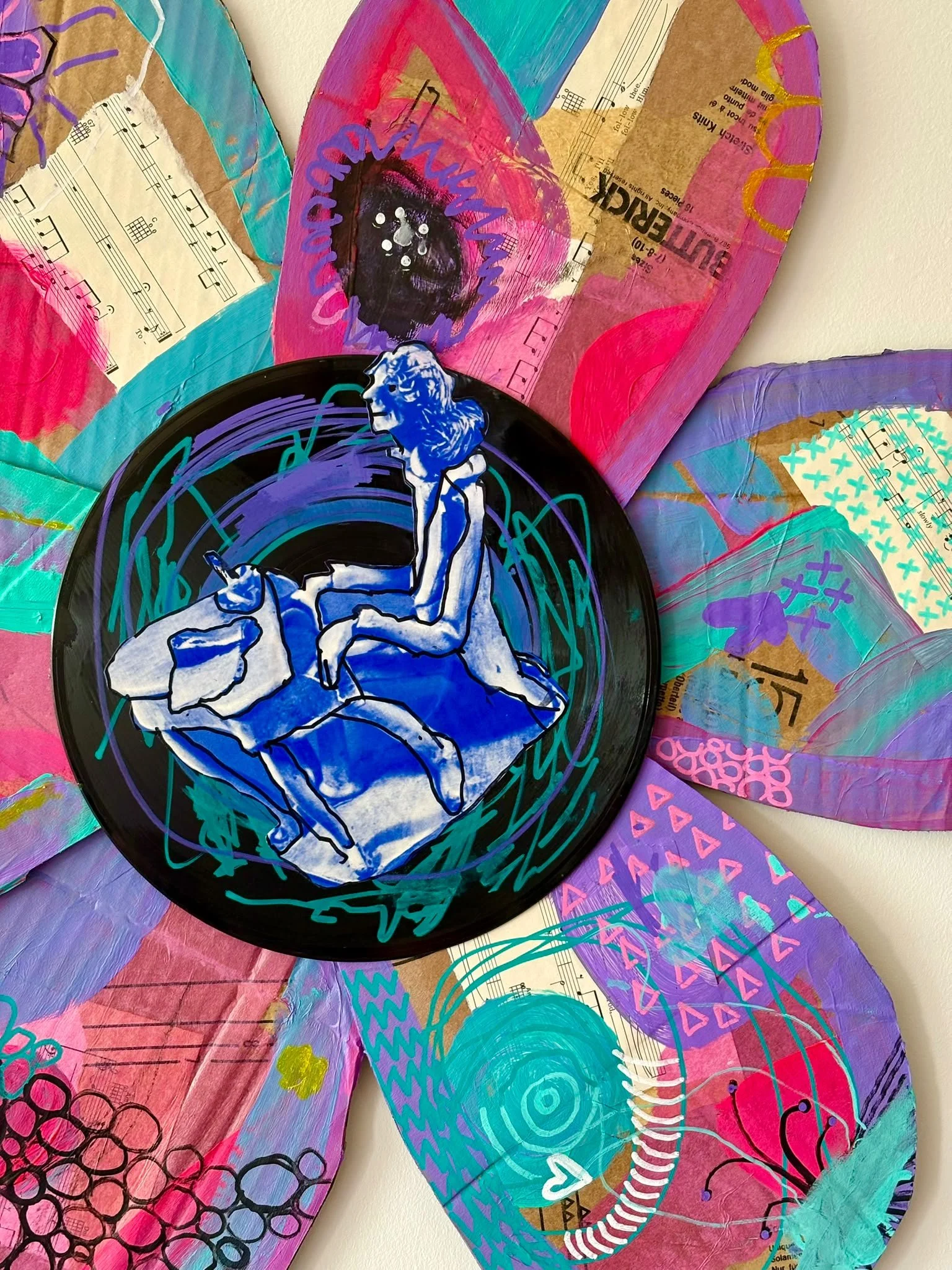 Colorful mixed media art piece featuring painted cardboard, sheet music, illustrations of lotion bottles, and a black record with a white and blue illustration of a seated woman reading. Bright colors of pink, purple, blue, and turquoise dominate the artwork.