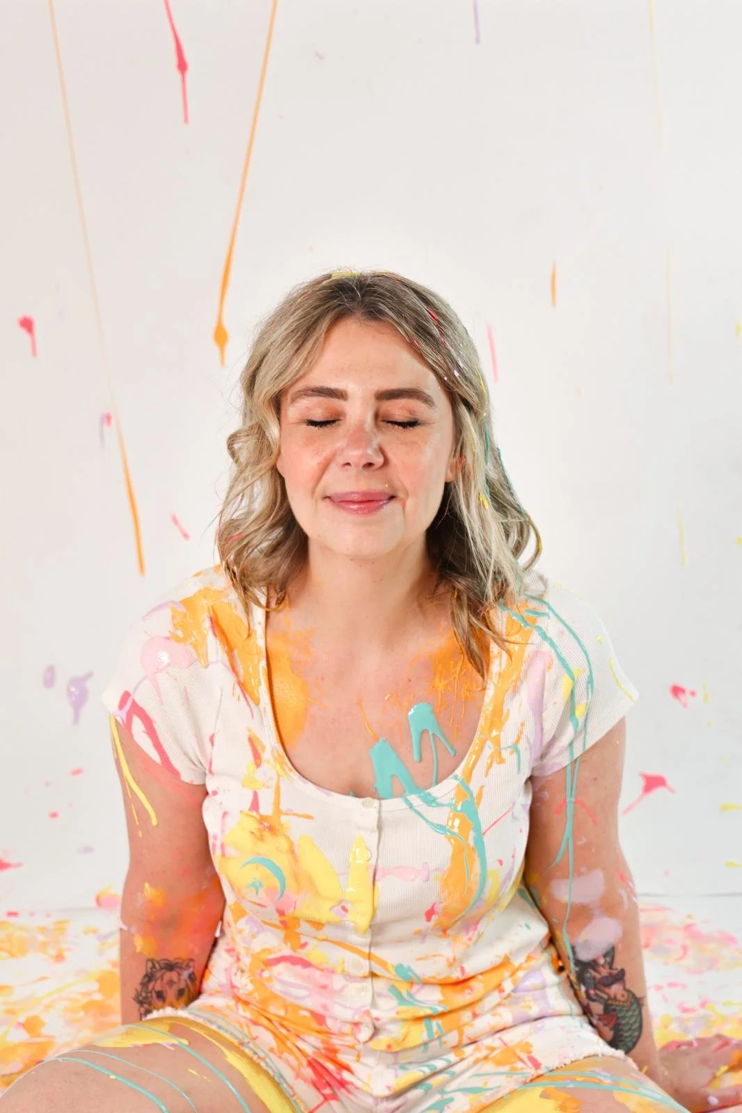 A woman with closed eyes, smiling softly, sitting with her legs crossed, covered in colorful paint splatters on her clothes and skin, with a white background decorated with hanging streamers.