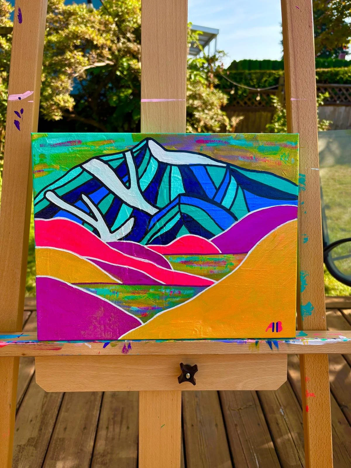 Colorful abstract painting of mountains with blue, green, pink, and yellow hues, displayed on an easel outdoors.