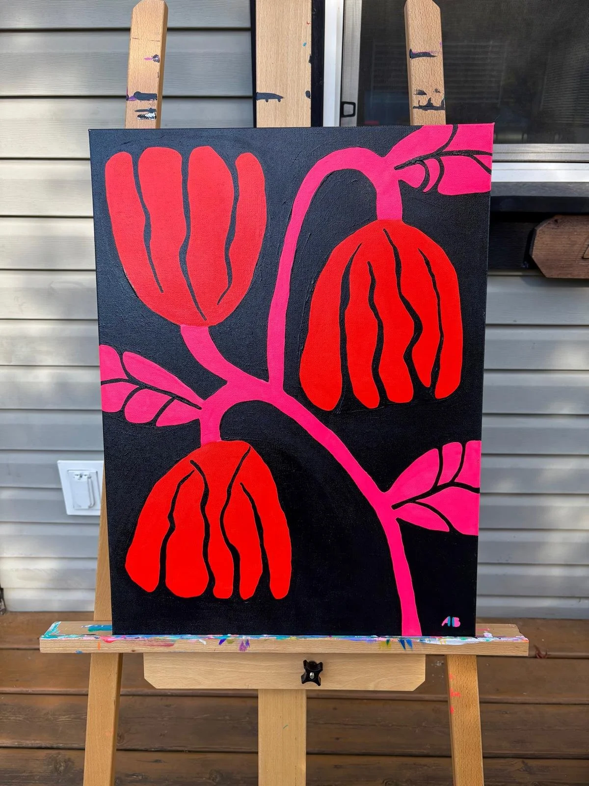 A painting of red, orange, and pink tulips with black outlines on a black background, displayed on an easel outside a house.