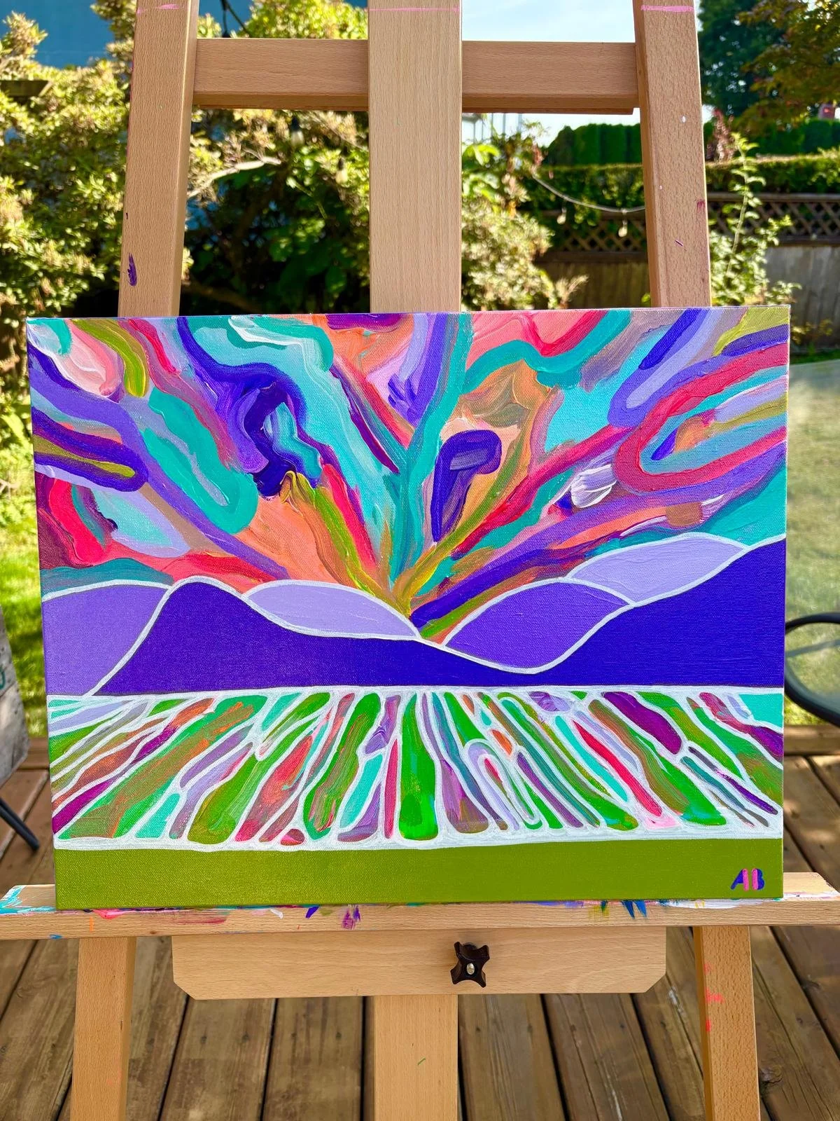 Colorful abstract painting on canvas, depicting a mountain with vibrant, swirling patterns resembling fireworks or bursts of energy, set outdoors on an easel in a garden.
