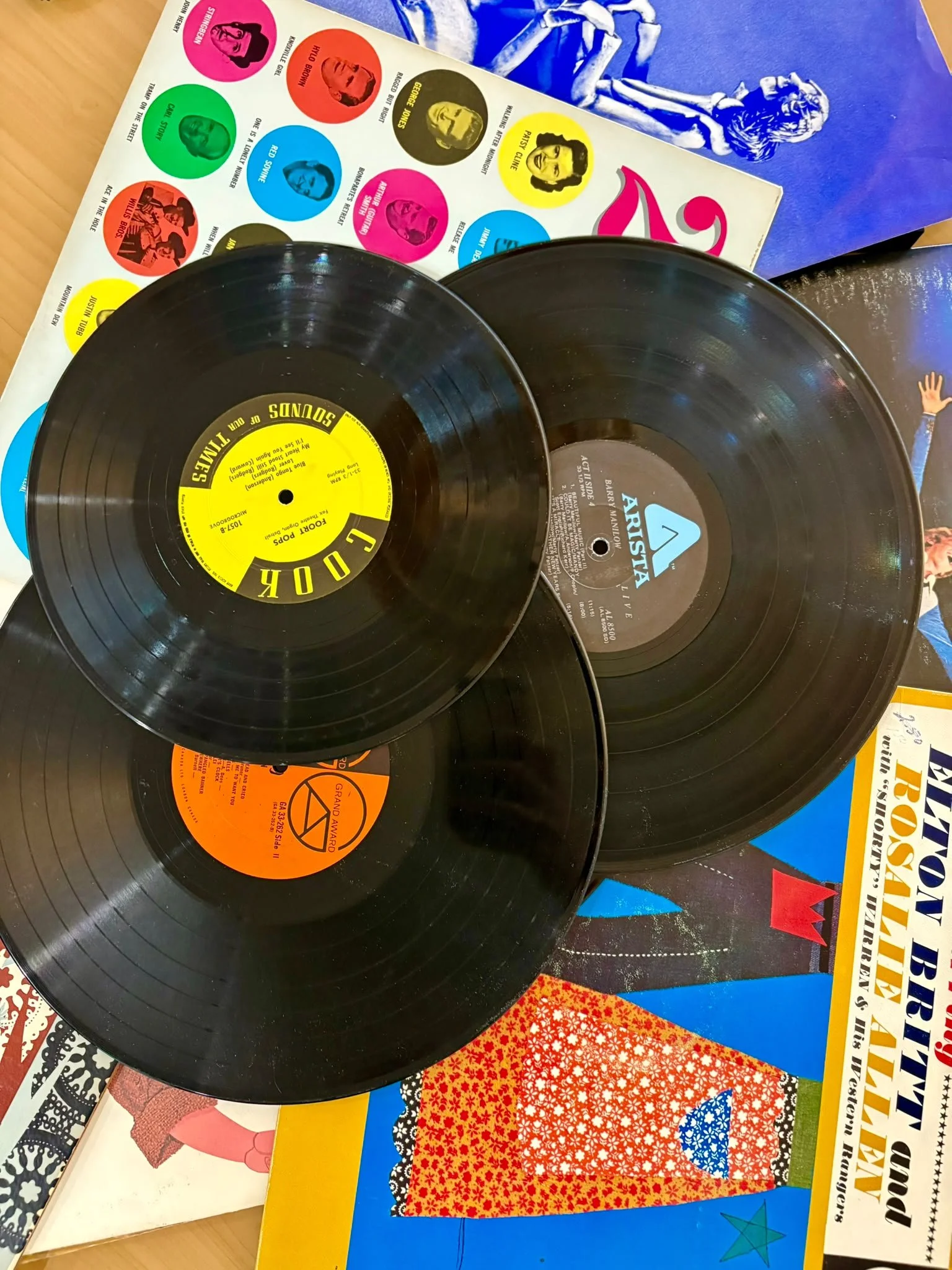 Two black vinyl records with yellow and gray labels on a colorful table covered with various posters and prints, including a sheet of multicolored circles with photographs and names of Donald Trump, George Jones, and other personalities.