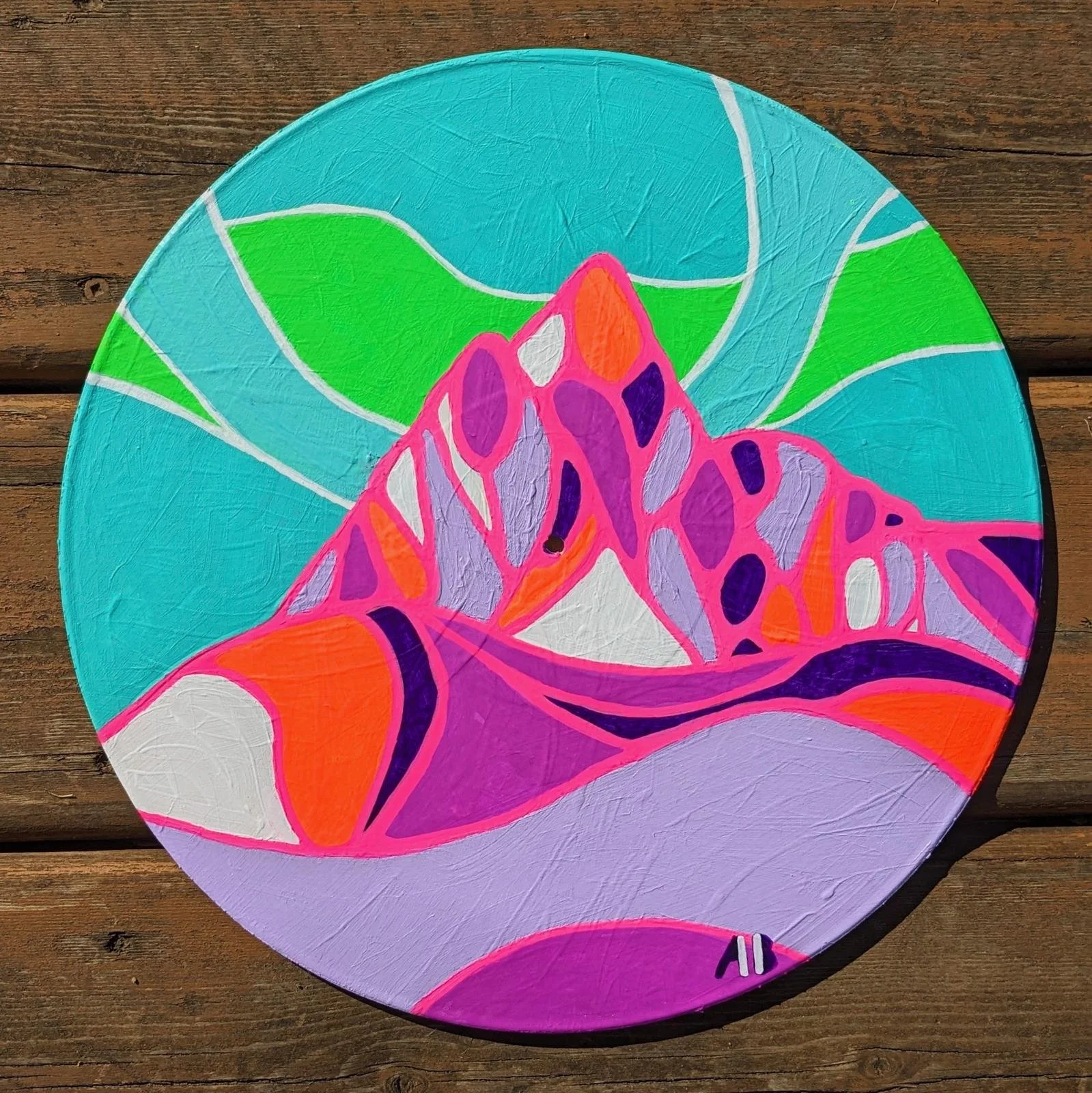 A colorful abstract painting on a round canvas features vibrant colors including pink, purple, orange, green, blue, and white. The design consists of various shapes and patterns, resembling organic forms or stained glass. The canvas is placed on a wo