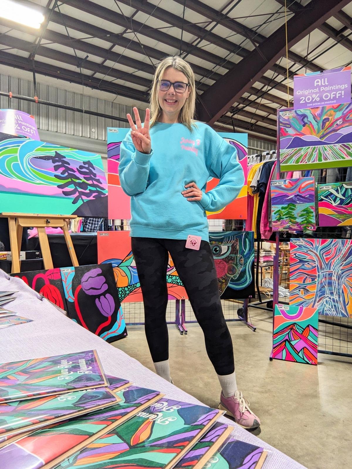 A woman standing at an art booth displaying colorful abstract landscape paintings. She is smiling and making a peace sign with her right hand.