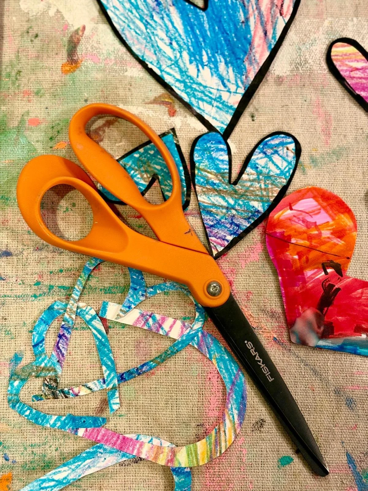 Craft supplies including cut-out paper hearts with colorful patterns, a pair of orange scissors, and a splattered canvas background.