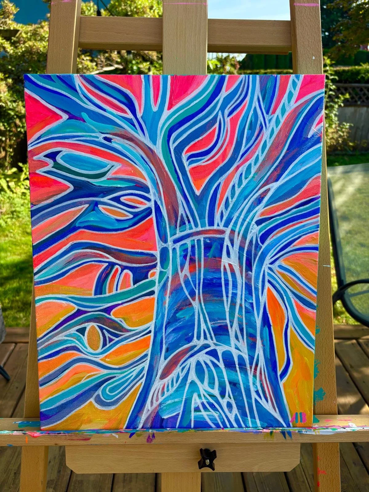 An abstract painting of a stylized tree with swirling blue, white, and purple lines on a bright orange and yellow background. The artwork is on a canvas mounted on an easel outdoors.
