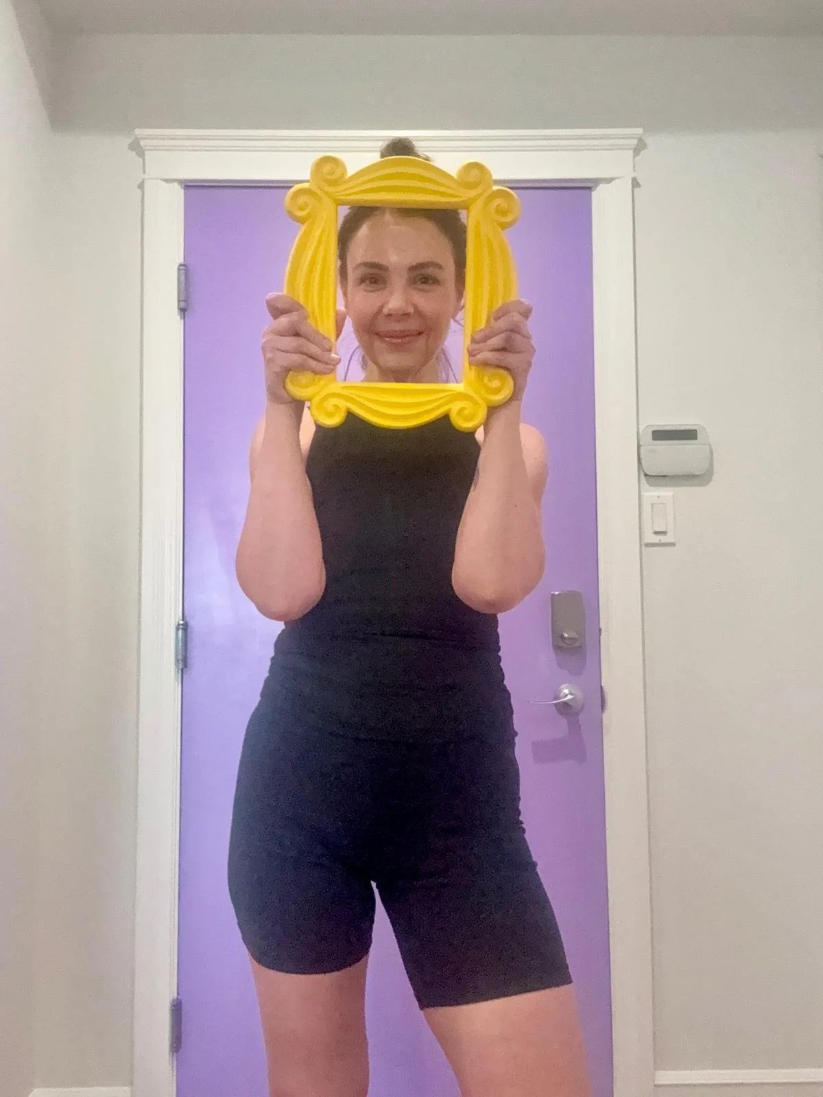 A woman taking a selfie in front of a purple door while holding a yellow decorative frame. She is wearing a black sleeveless top and matching black shorts, and has her hair tied up.