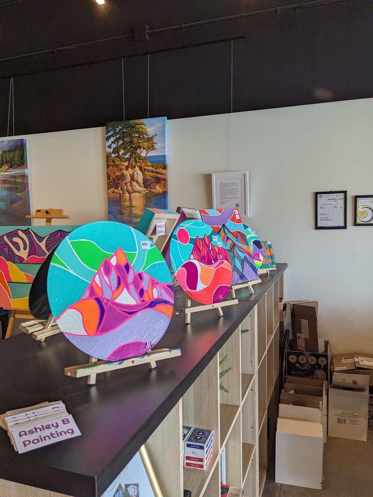 Colorful landscape paintings and circular art pieces on display at an art gallery or store. The artworks feature vibrant colors and abstract mountain scenes. Framed documents are hung on the white wall behind the display.
