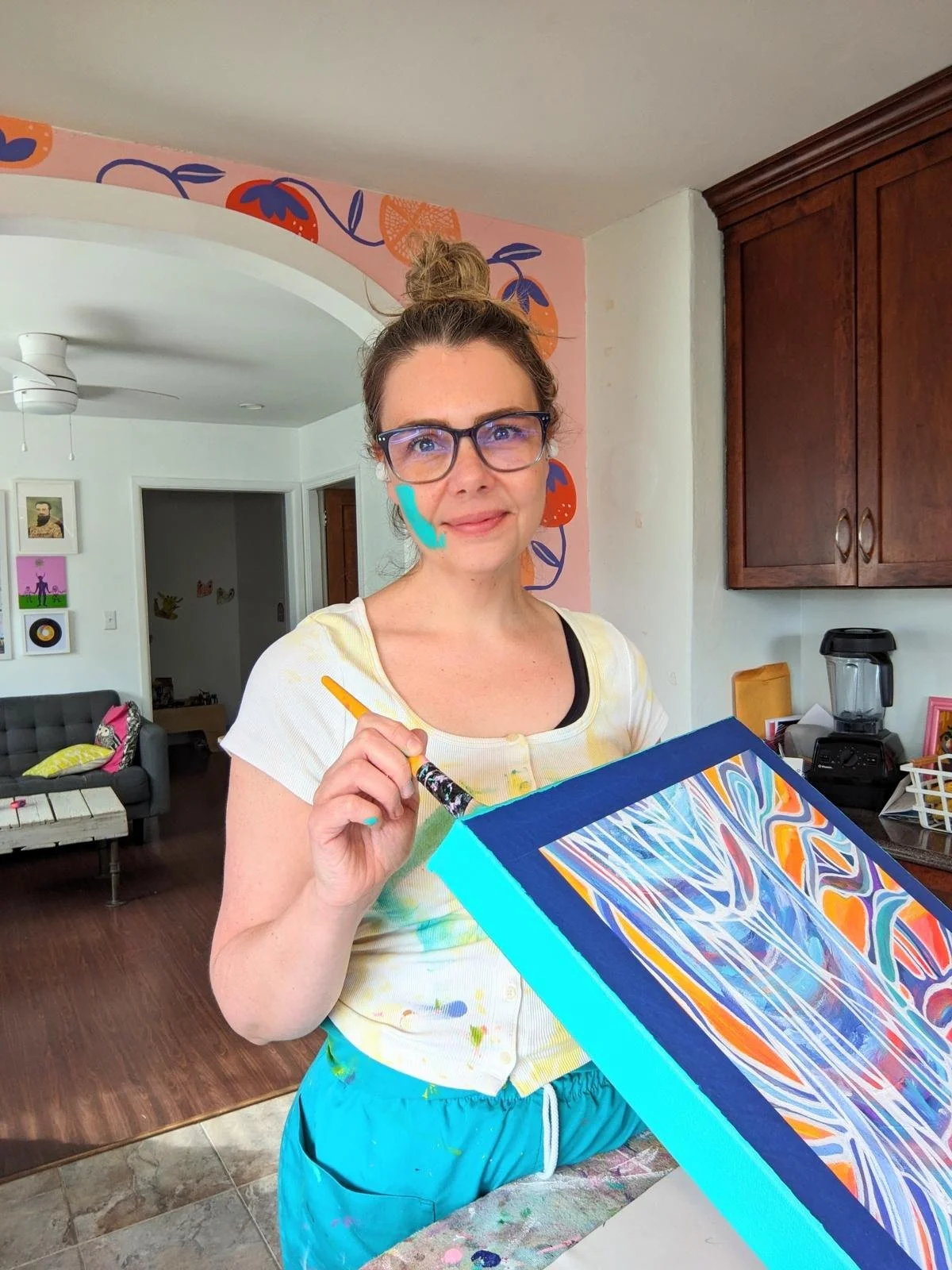 Woman with glasses and bun hairstyle holding a paintbrush and a colorful, abstract painting in a kitchen.