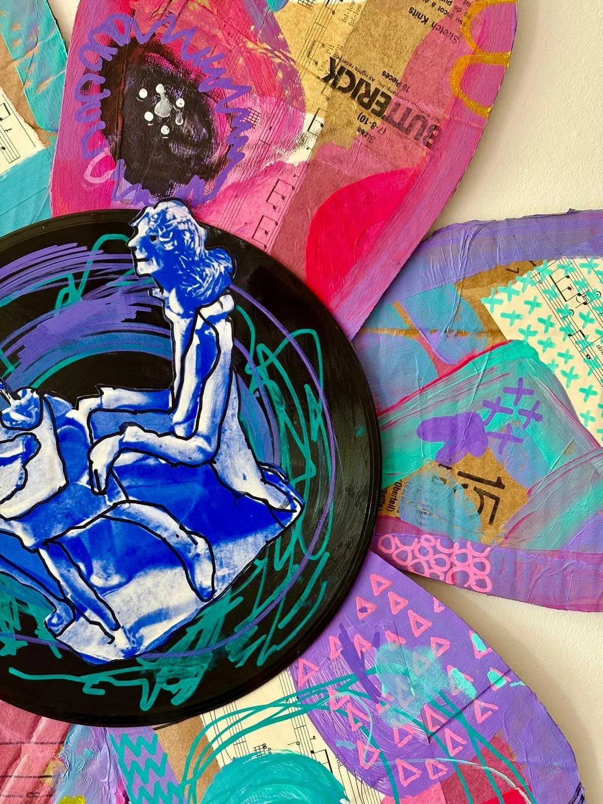A black vinyl record with a blue and white drawing of a person sitting cross-legged is placed over colorful abstract artwork and collaged paper with various shapes, scribbles, and patterns.