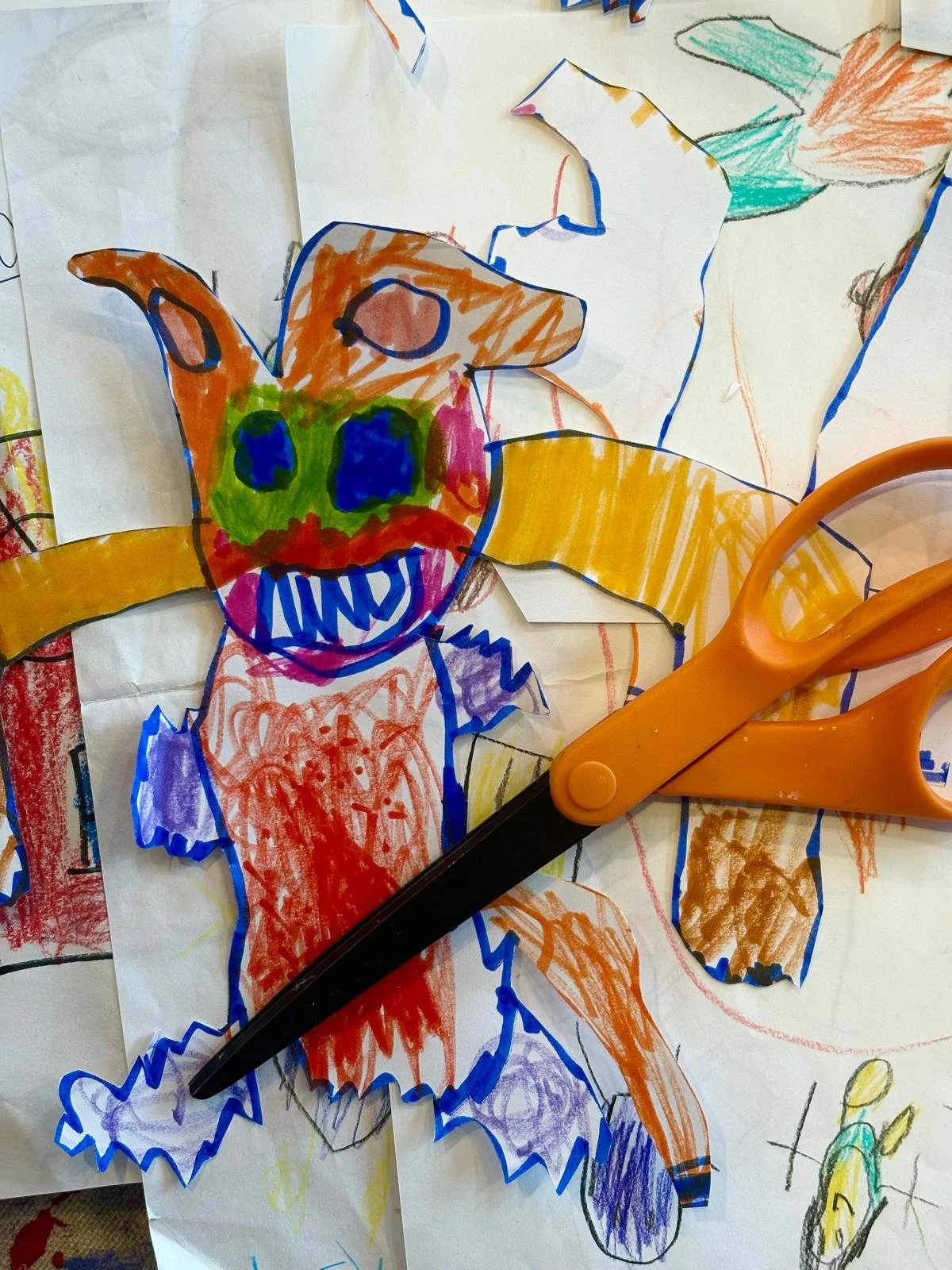 Child's drawing of a colorful clown with orange hair, green face, blue eyes, and a big, toothy smile. The clown has large orange ears and wears a multicolored outfit. An orange pair of scissors is also visible on the paper.