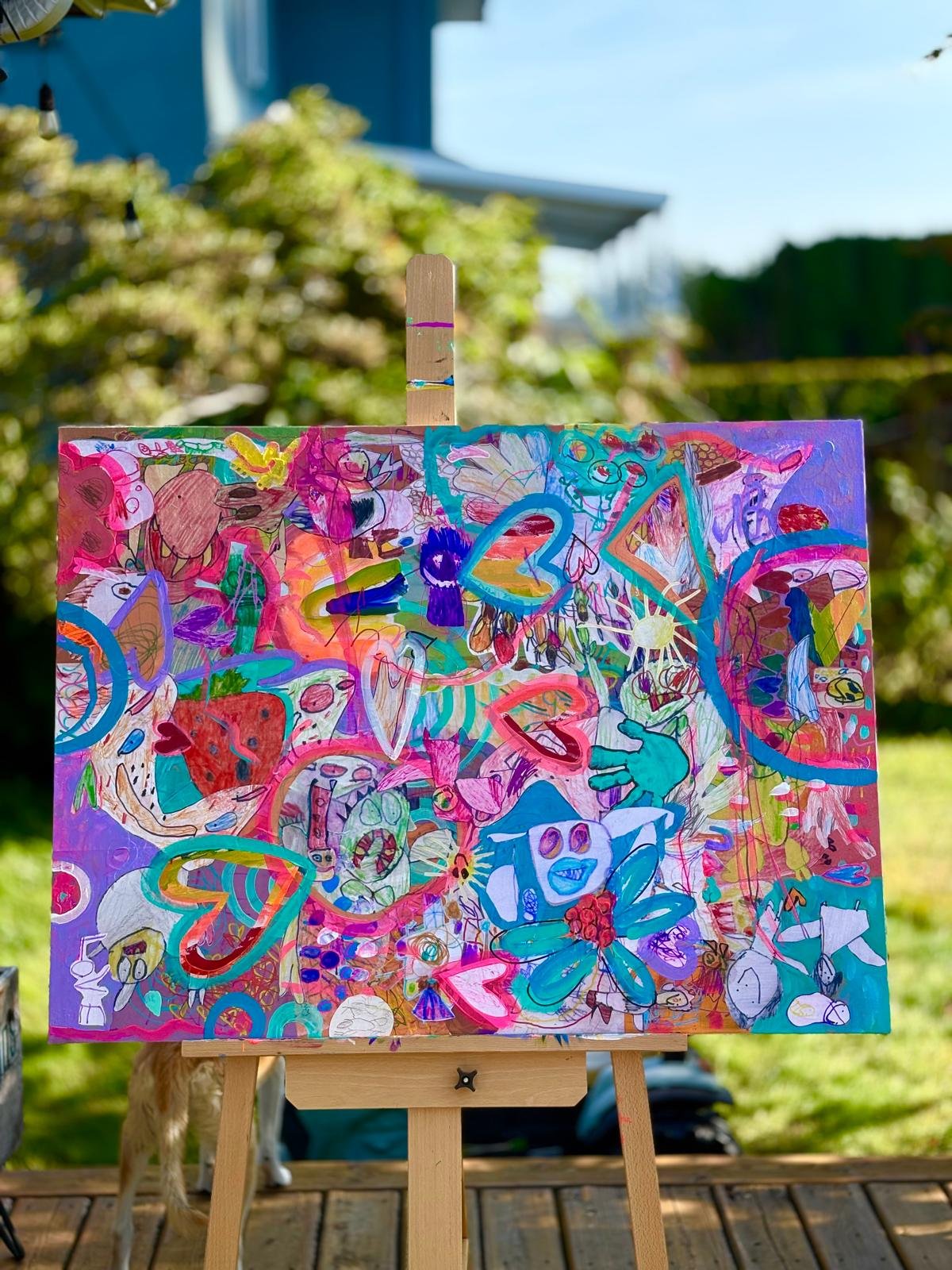 Colorful abstract artwork on a canvas with various shapes, patterns, and doodles, mounted on an easel in an outdoor setting with greenery and a house visible in the background.