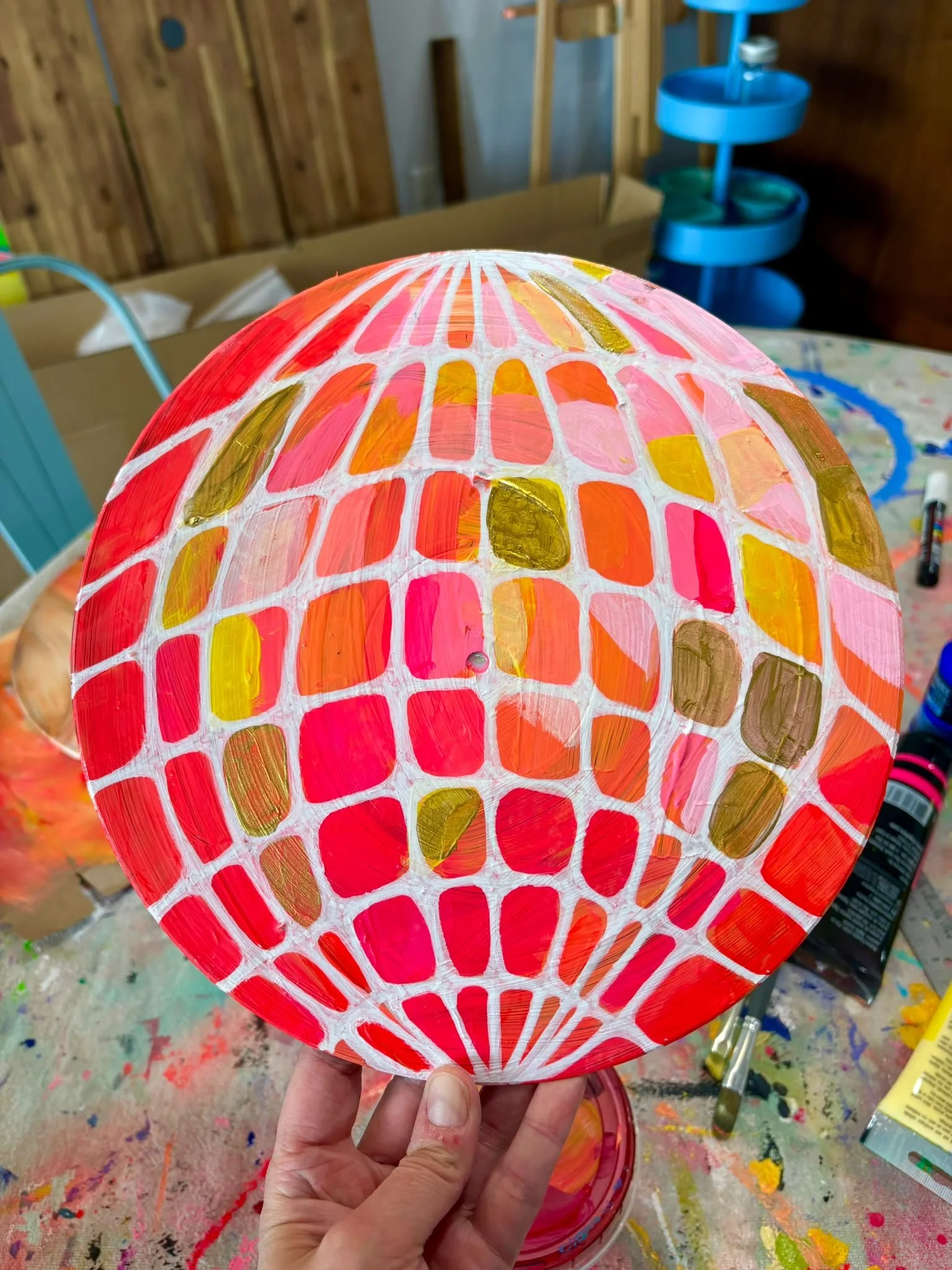 Close-up of a decorated paper lantern featuring vibrant pink, orange, yellow, and gold patterns with a web-like white outline, held by hand above a cluttered, colorful workspace.