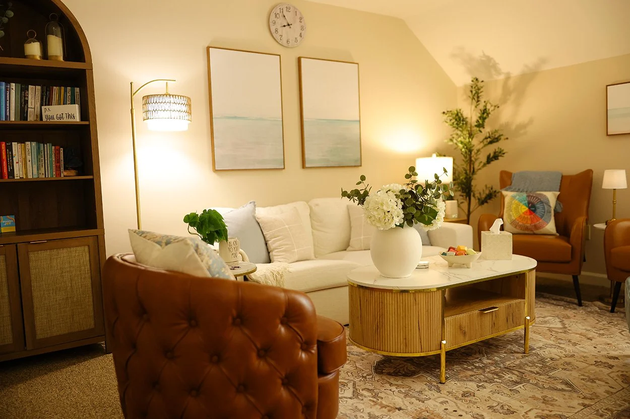 Cozy living room decorated with paintings, a white sofa, brown armchairs, a marble-topped coffee table with flowers, and various lamps and plants.