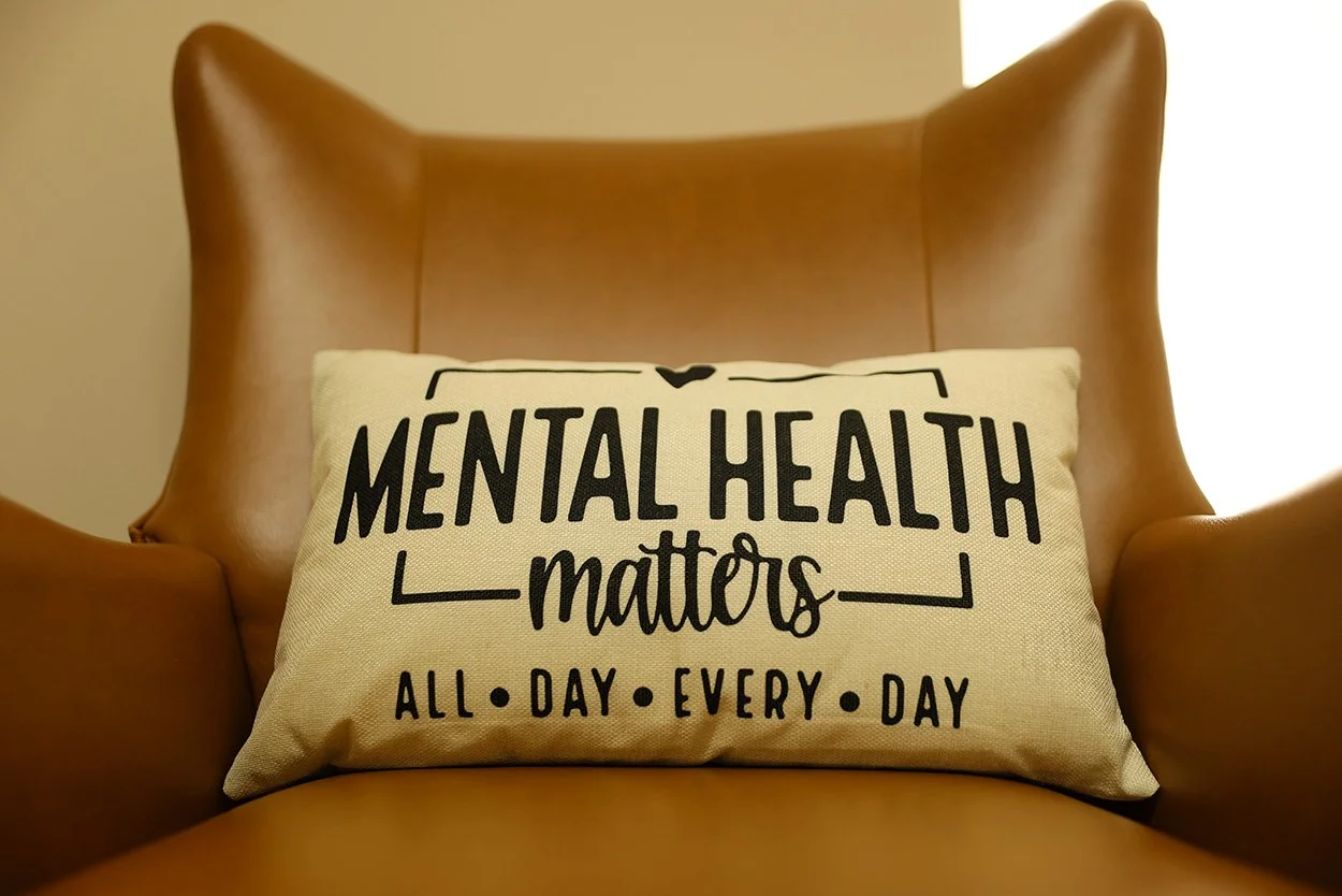 Pillow with text 'MENTAL HEALTH matters ALL DAY EVERY DAY' on a brown leather armchair.