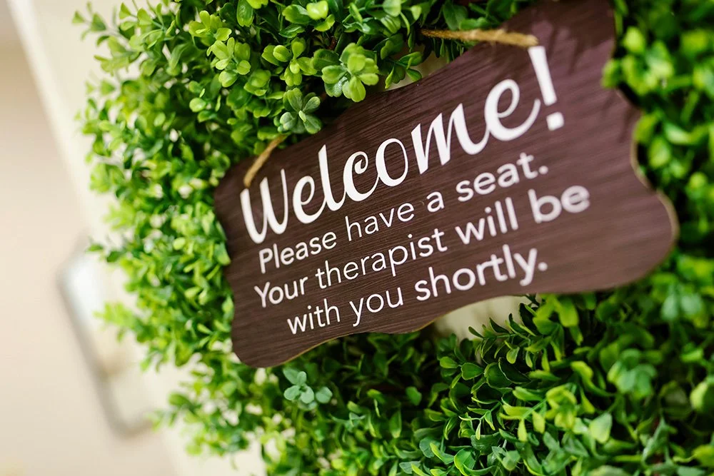 Wooden welcome sign among green foliage with white text that says, "Welcome! Please have a seat. Your therapist will be with you shortly."