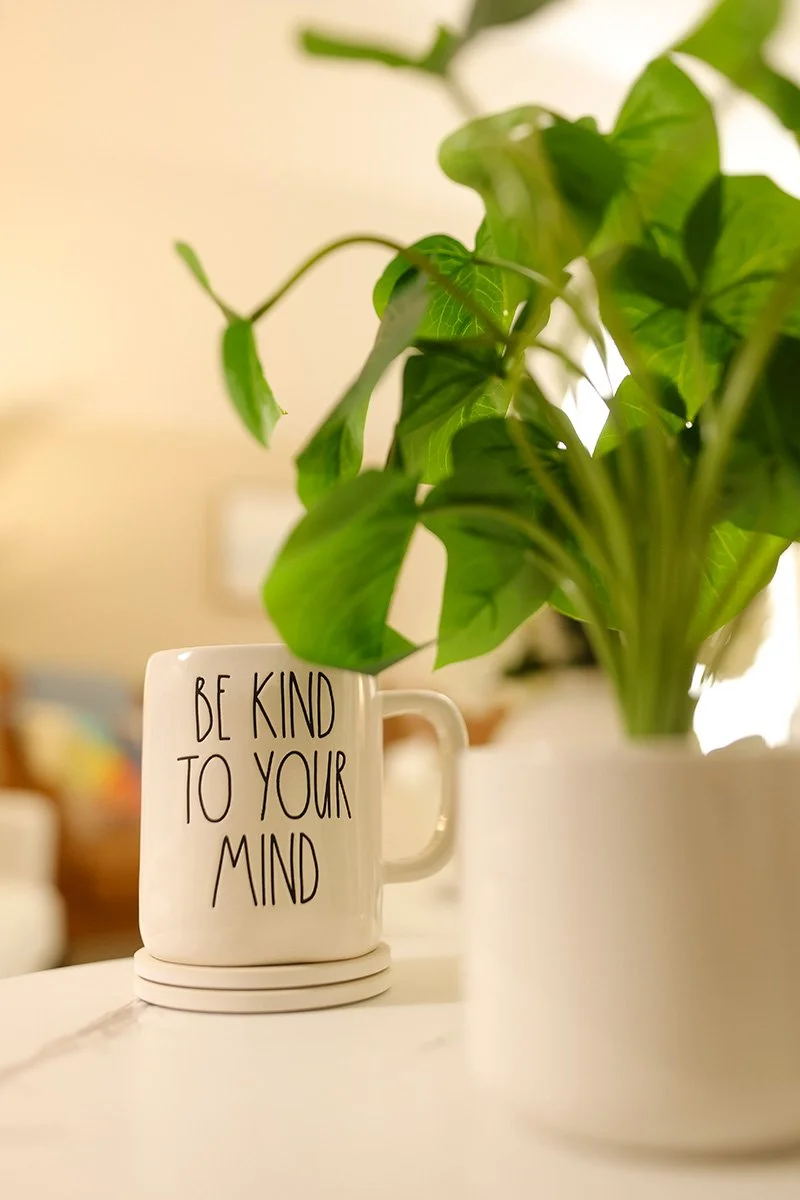 A white mug with the words 'Be kind to your mind' written on it, placed on a table next to a potted green plant.