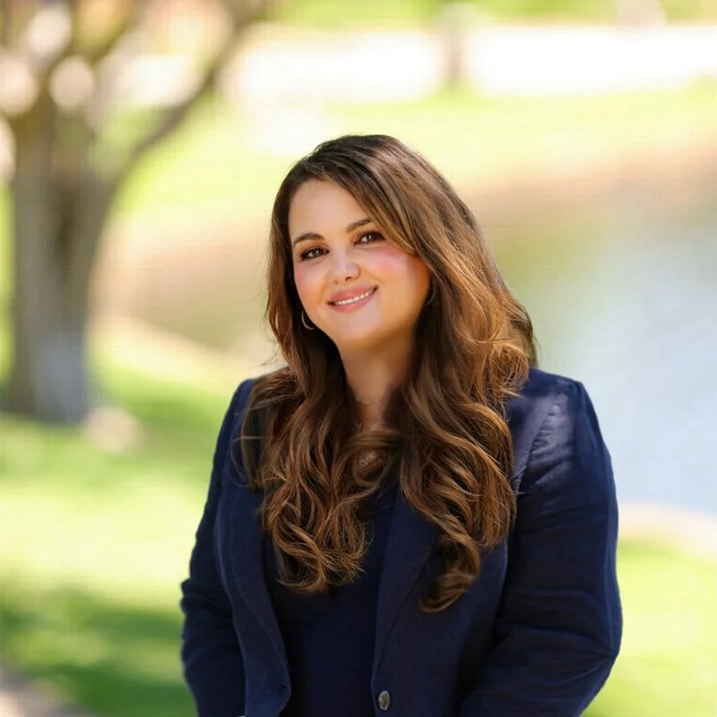 A woman with long, wavy brown hair wearing a navy blazer, smiling outdoors with trees and water in the background.