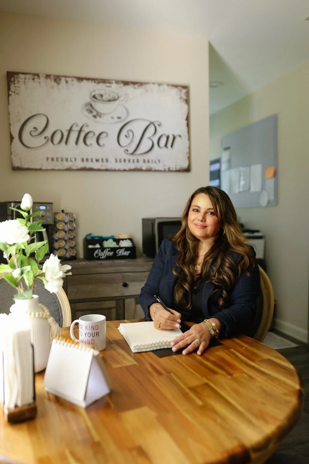 A woman with long, wavy brown hair sits at a wooden table in a coffee shop, smiling at the camera. On the table are a white mug with red text, a small potted plant with white flowers, and notebooks. Behind her, a sign reads "Coffee Bar," and the background includes a coffee machine and other equipment.