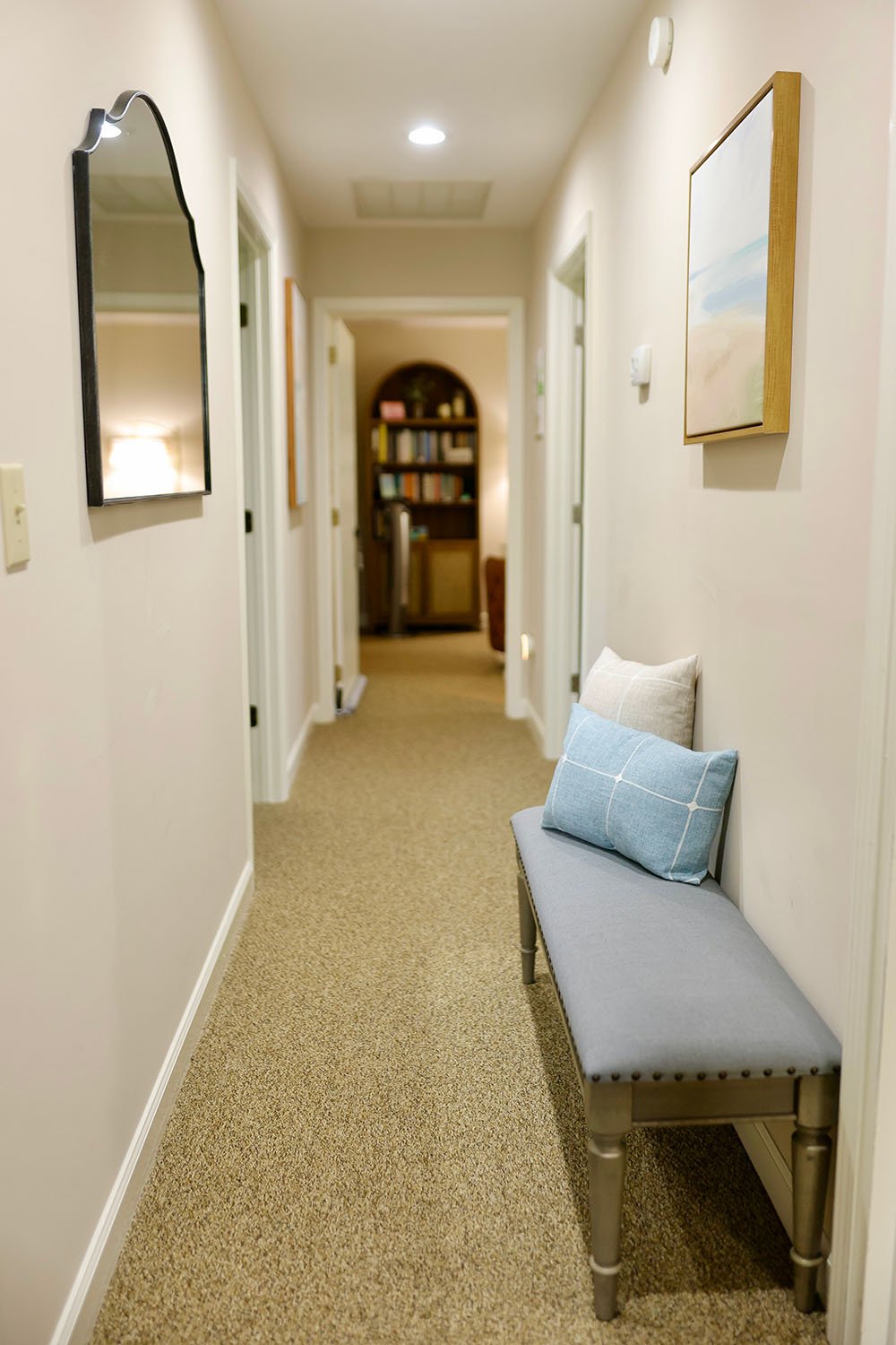 Hallway with a bench and pillows, framed artworks on the wall, and a bookshelf in the background.