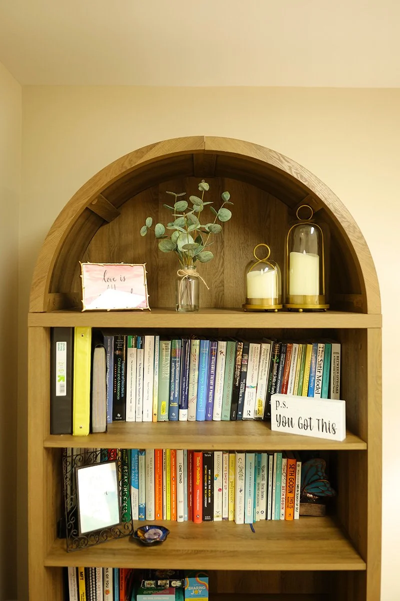 A wooden bookshelf with an arched top containing various books, a small mirror, and decorative candles. On top, there's a glass vase with eucalyptus branches, a framed quote that says 'love is all you need,' and two glass-covered candles.