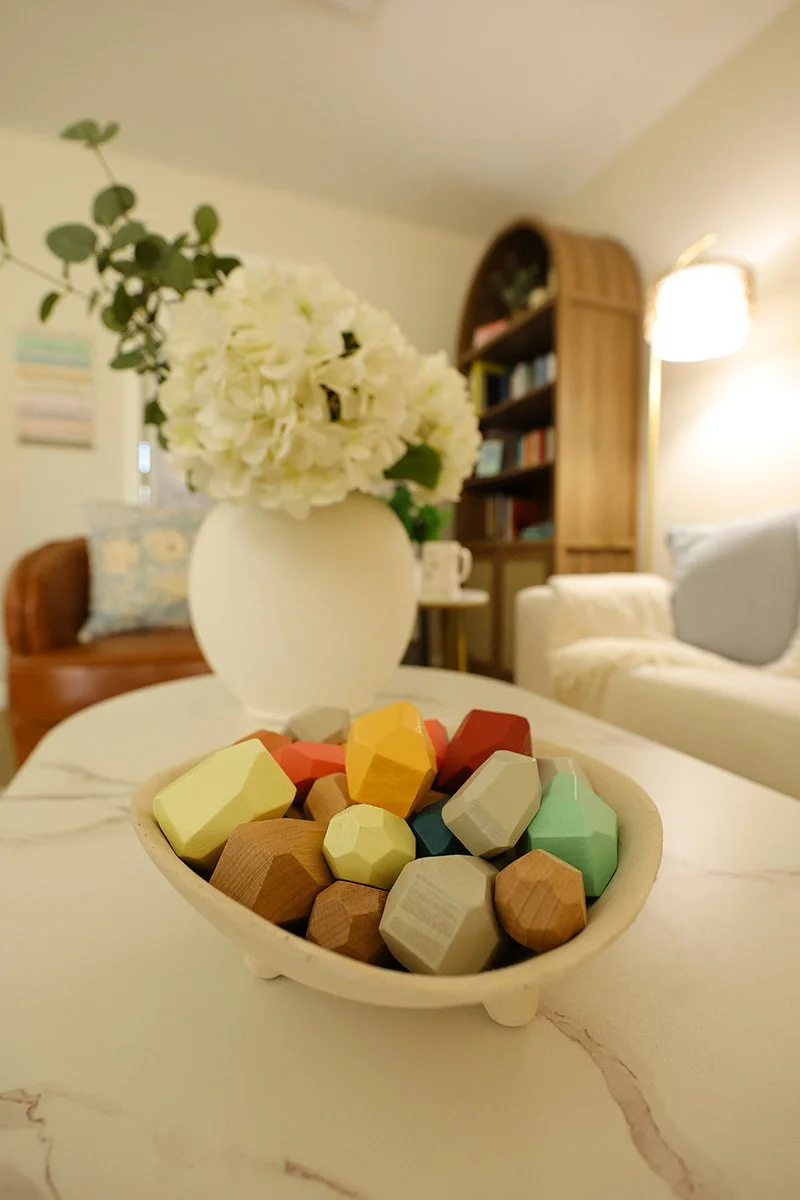A white marble table with a bowl of colorful geometric wooden toy blocks in the foreground, and a white vase with white flowers and green leaves in the background, in a cozy living room with bookshelves and soft lighting.