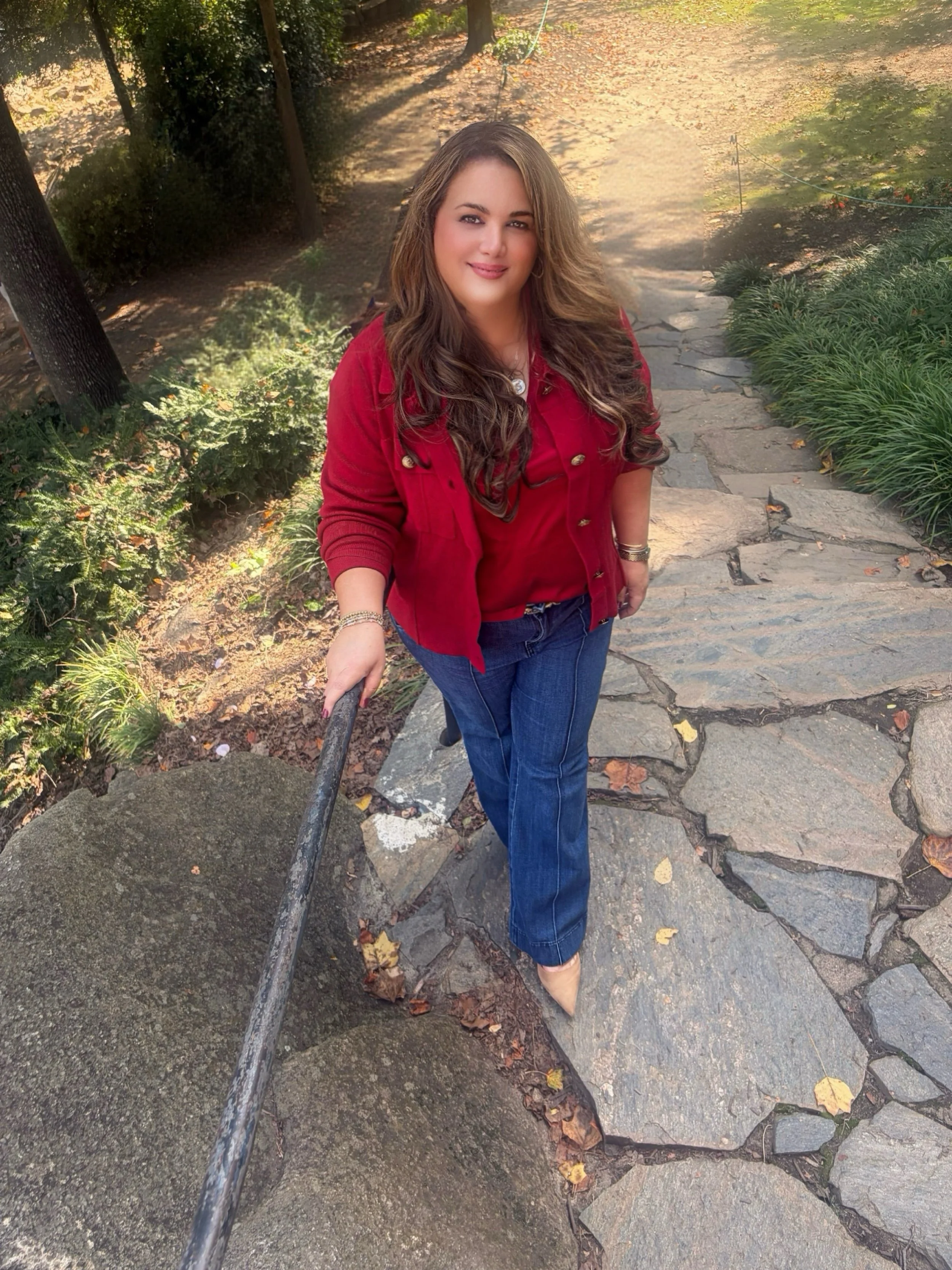 A woman with long, wavy brown hair, wearing a red jacket and blue jeans, taking a selfie on a stone pathway in a park surrounded by trees and greenery.