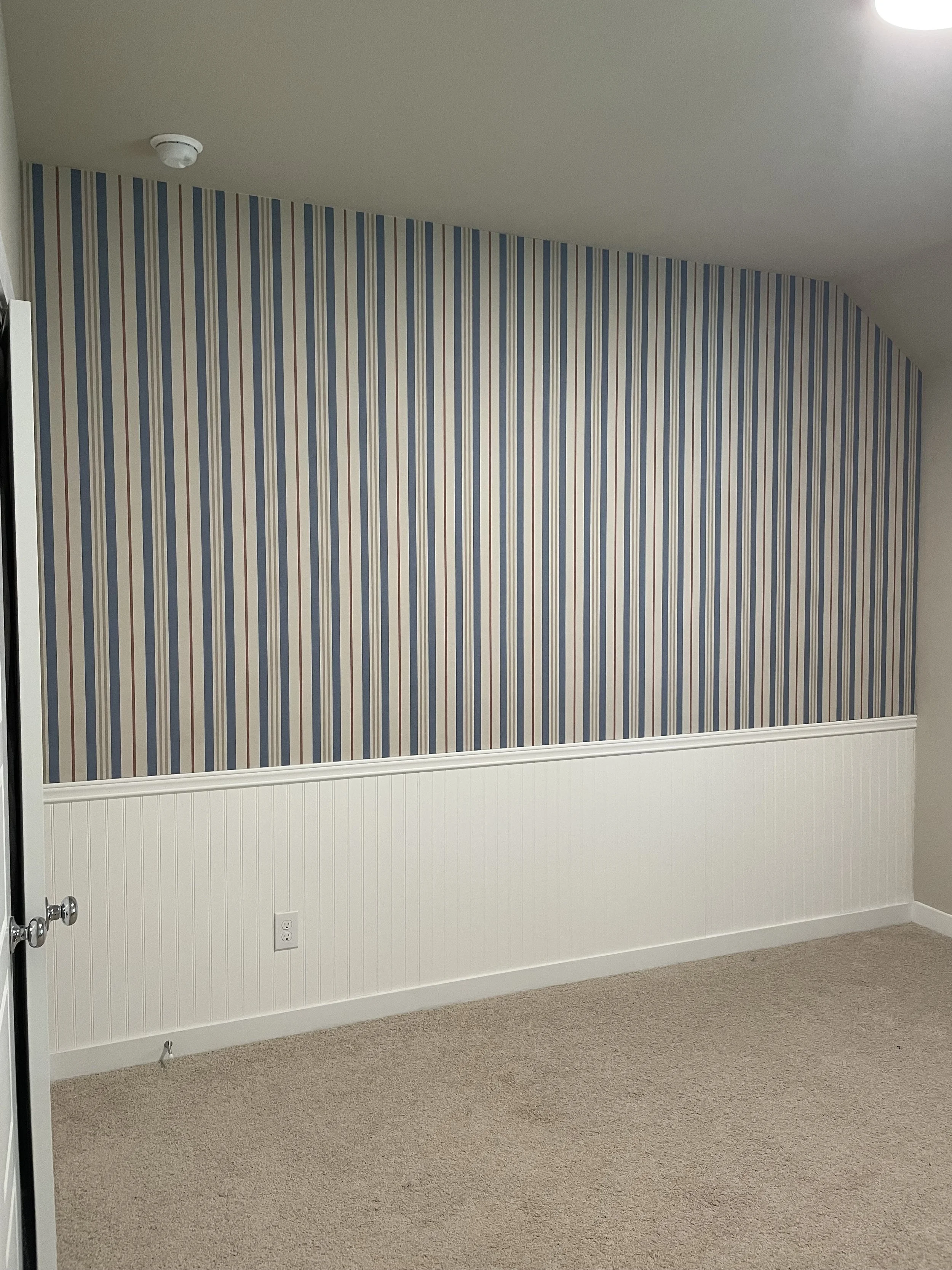 Empty room with striped wallpaper on one wall, white wainscoting on lower wall, beige carpet, ceiling light, and a smoke detector.