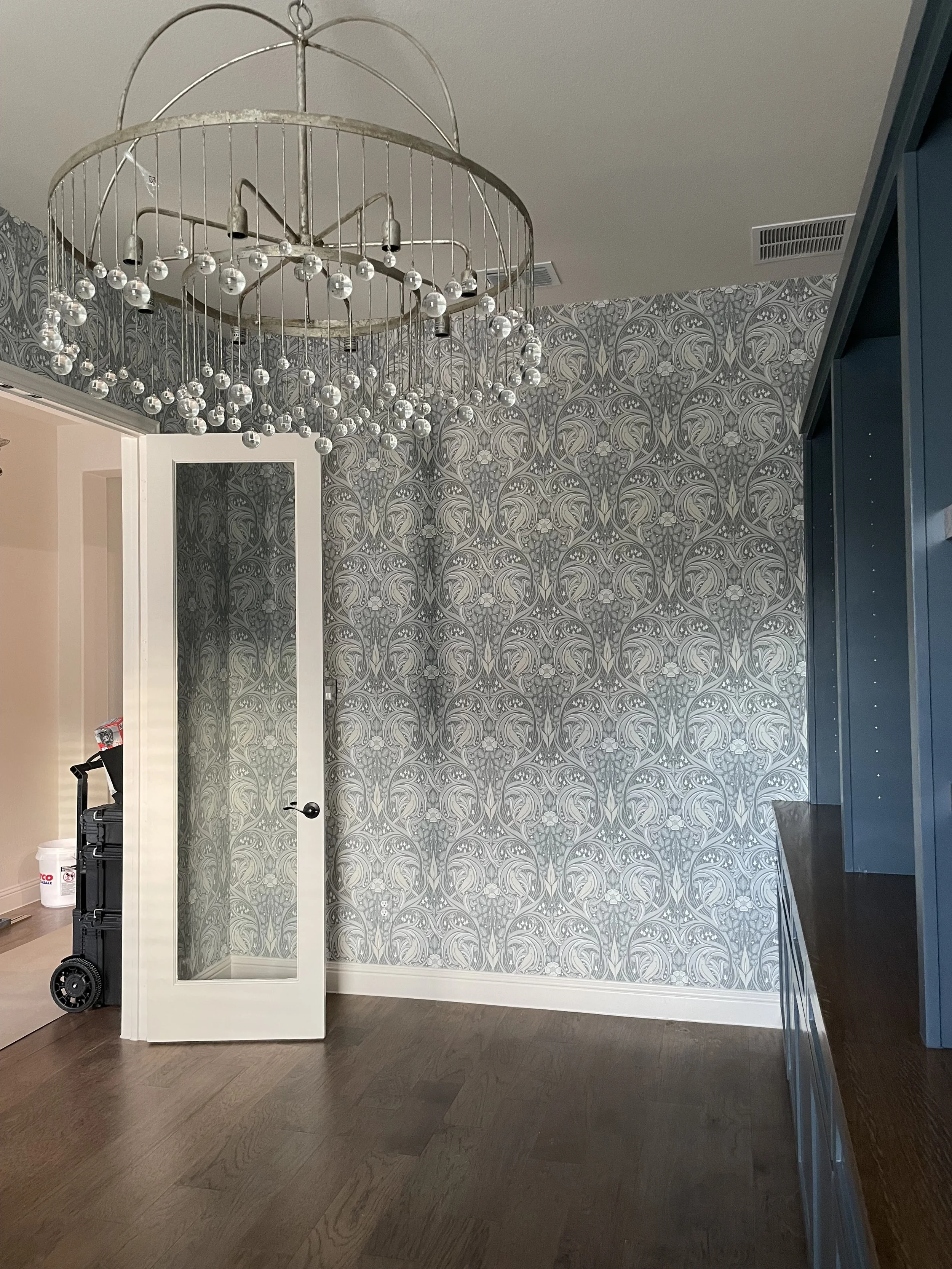 Interior view of a room with blue patterned wallpaper, a white door with a glass pane, a dark blue cabinet, and a modern chandelier with glass orbs. There is a black luggage cart with some items on it and a white bucket placed on the floor near the door.