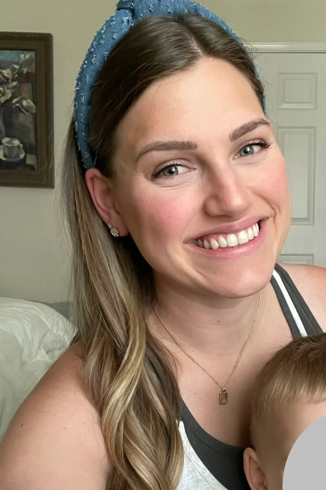 A smiling woman with long wavy light brown hair wearing earrings, a necklace, a blue headband, and a black and white tank top, indoors.