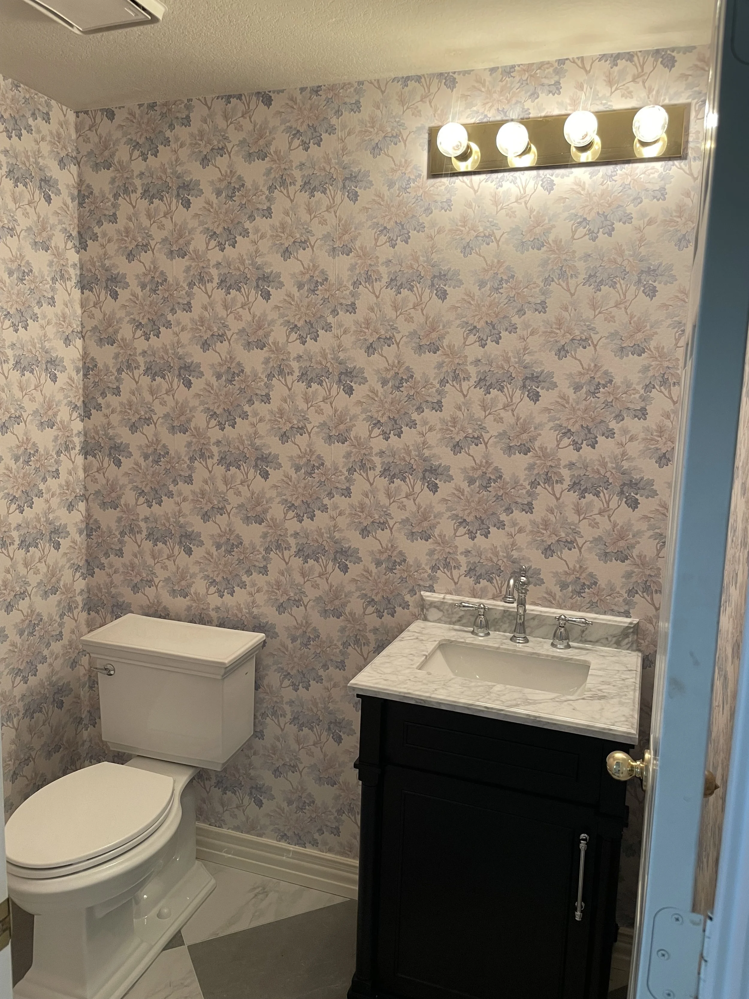 A small bathroom with floral wallpaper, a toilet, and a dark wooden vanity with a marble countertop and sink. There is a four-bulb light fixture above the mirror and an air vent on the ceiling.