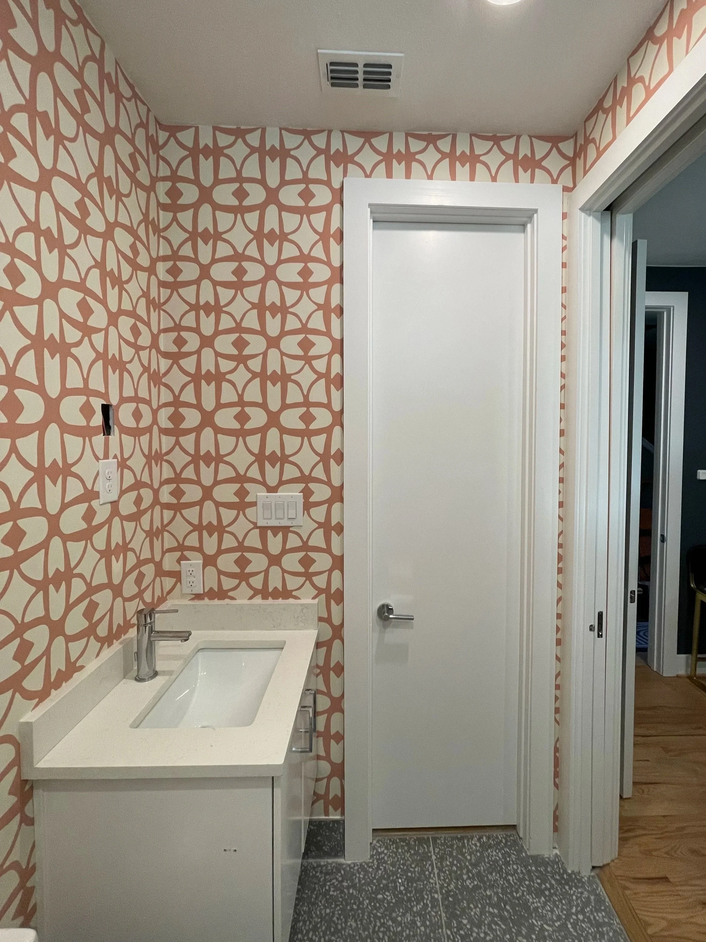 Bathroom with pink patterned wallpaper, white cabinet under a sink, white door with a silver handle, gray and black speckled tile floor, and a view into a hallway or other room.