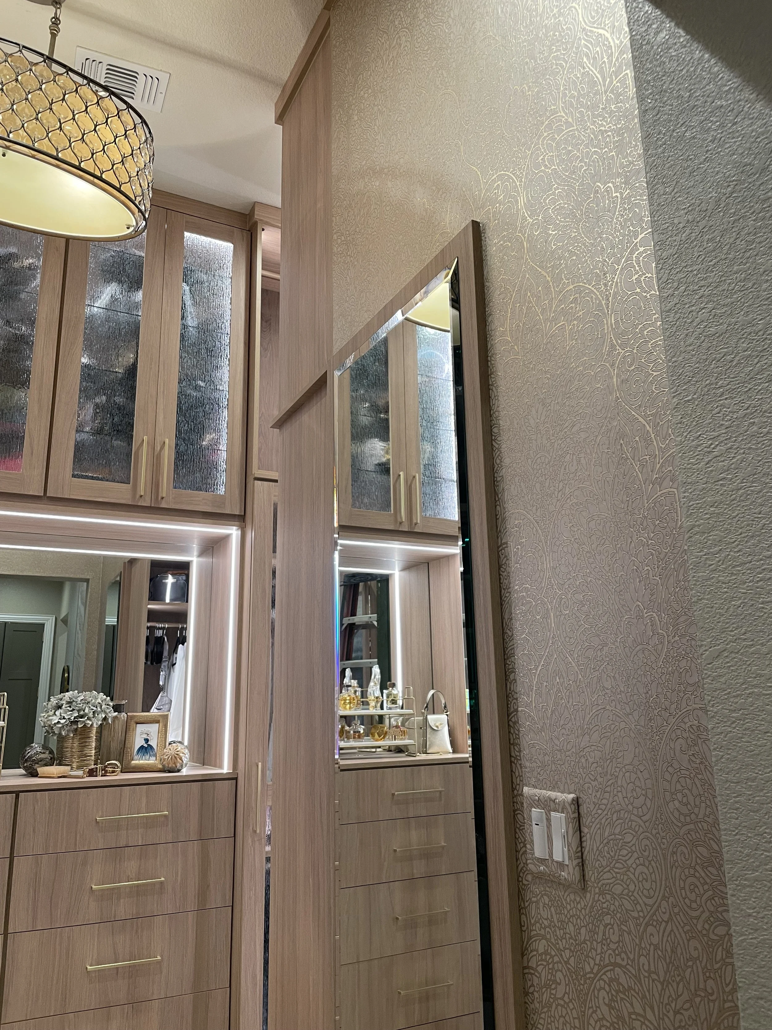 A modern wooden vanity with a mirror and gold handles, decorated with flowers and framed photos, with a textured wallpaper and a wall outlet below.