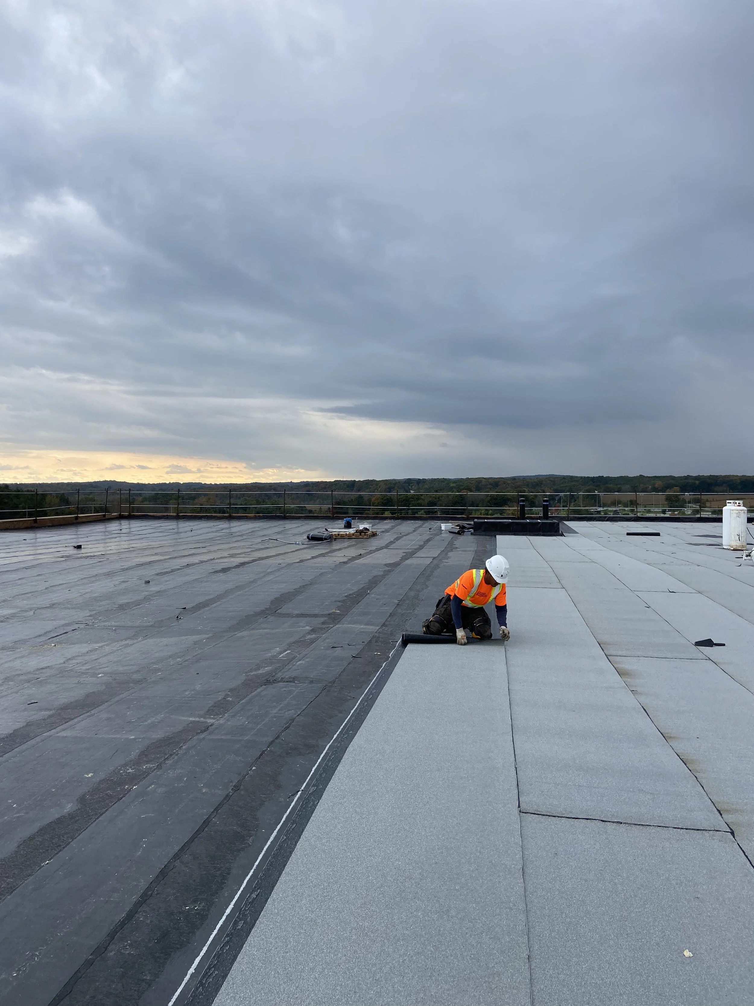A person wearing an orange safety vest and white hard hat is installing or repairing roofing materials on a flat rooftop on a cloudy day. The sky appears overcast with dark clouds, and there are some distant trees and a horizon.