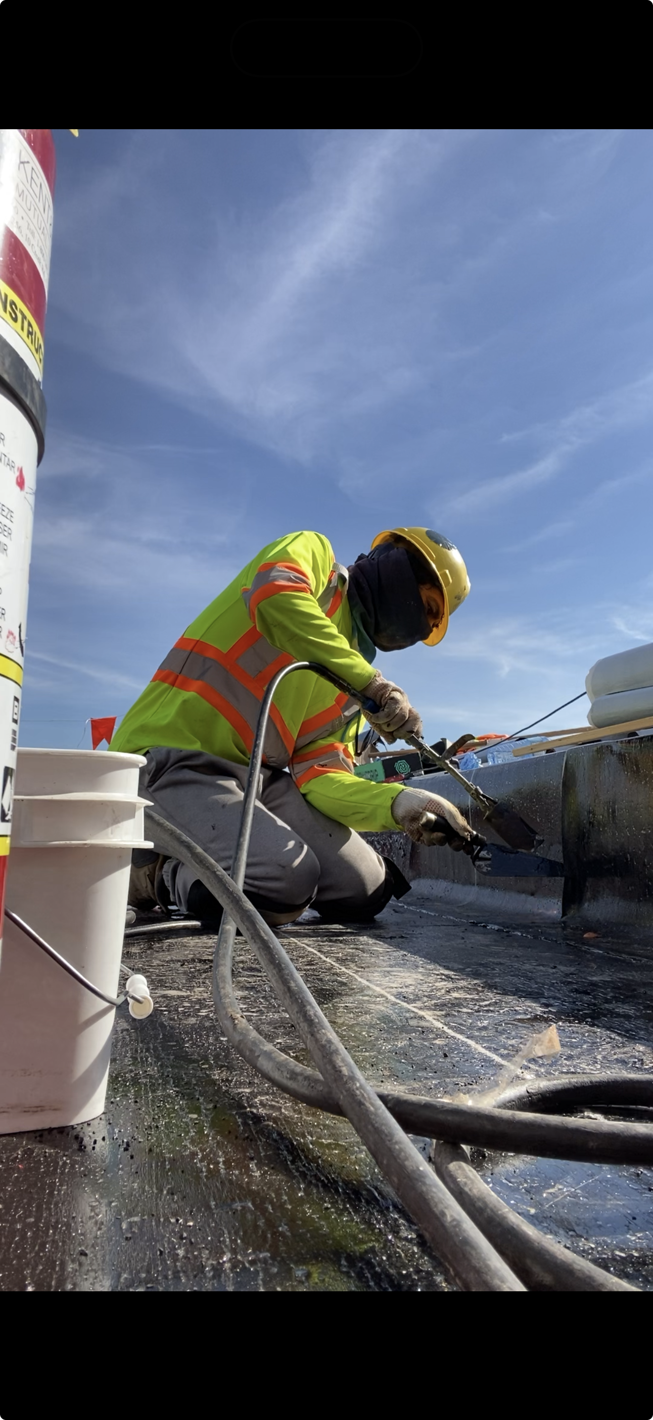 A construction worker wearing a yellow helmet, a high-visibility green jacket with orange stripes, black mask, and gloves is working on a boat or similar structure outdoors under a blue sky with few clouds. There are tools, cords, and buckets nearby, and the worker is focused on the task.