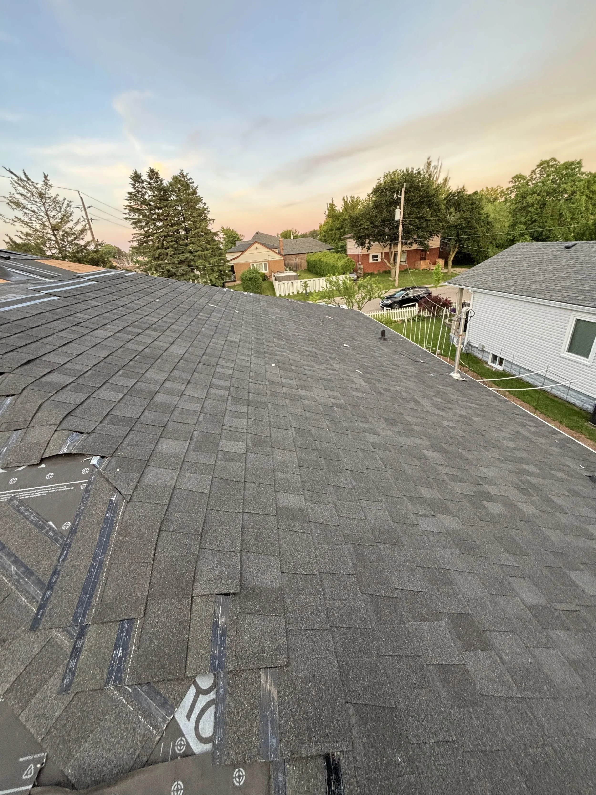View of residential neighborhood rooftops with asphalt shingles, trees, houses, parked car, and utility pole at sunset.