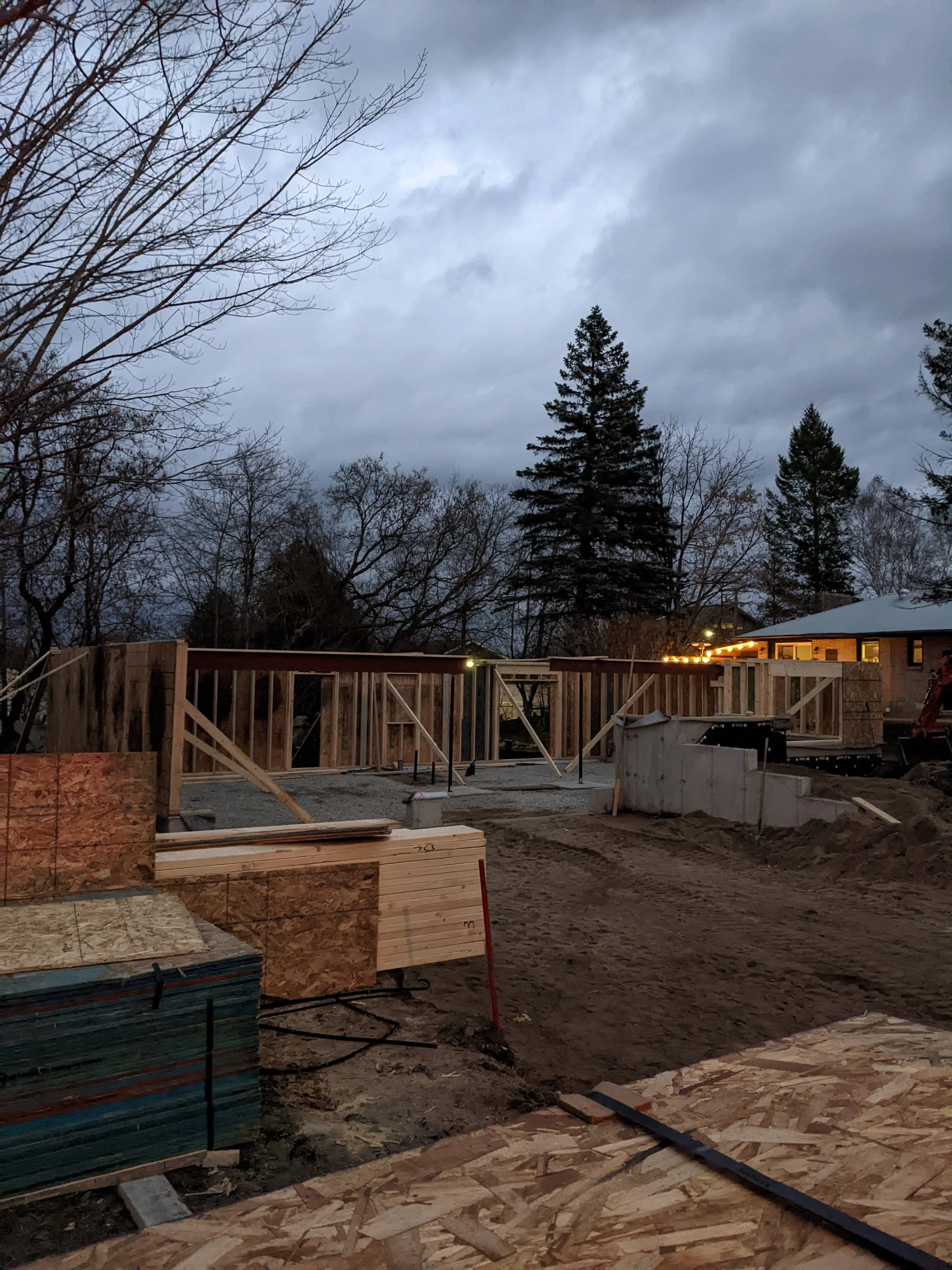 A construction site with wooden framing and partially completed foundation, under an overcast sky. Trees and a house with string lights are visible in the background.