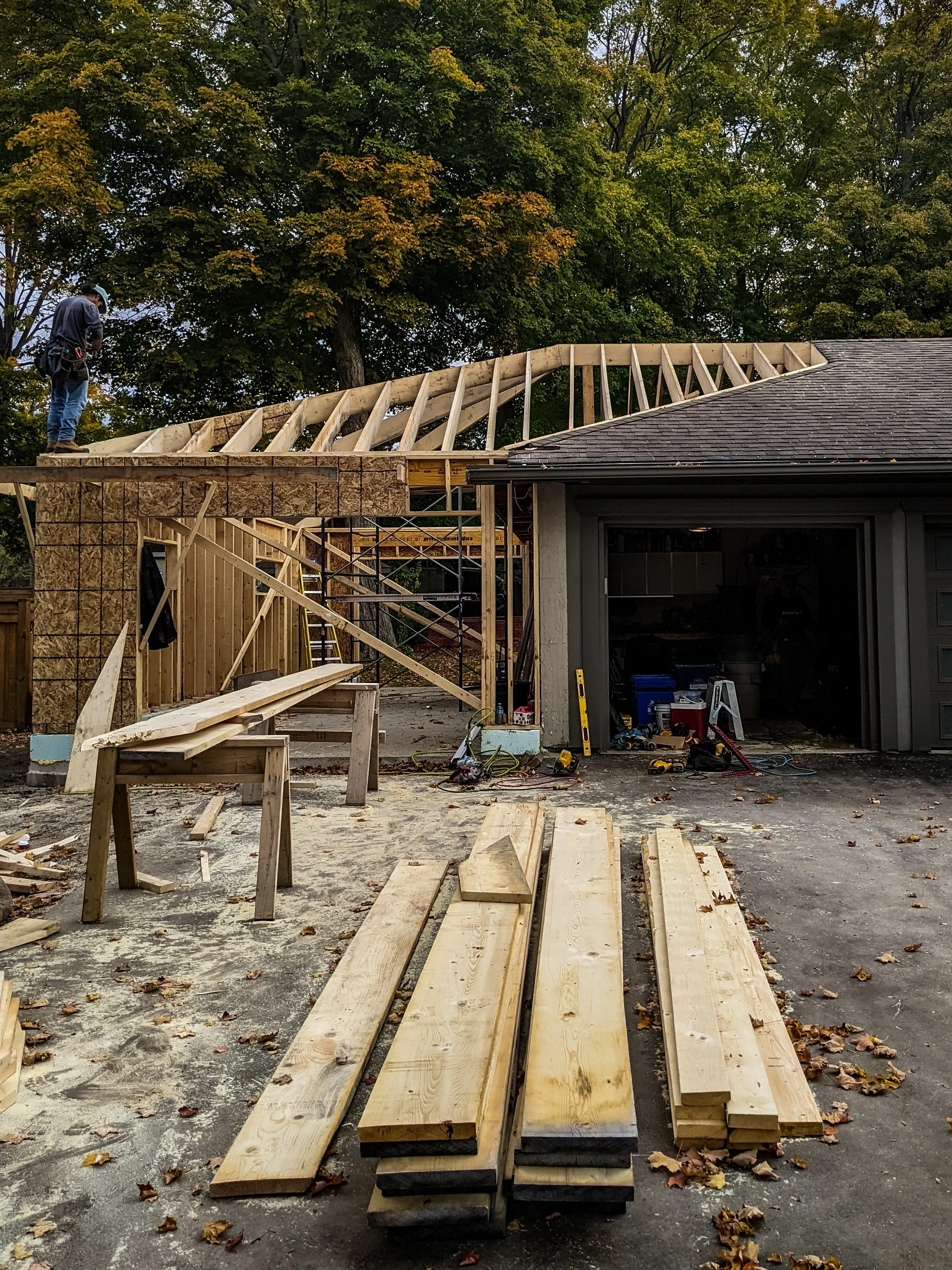 Construction site with a partially built roof and wooden beams, a worker standing on the roof framework, construction tools and wood planks on the ground, and trees in the background.